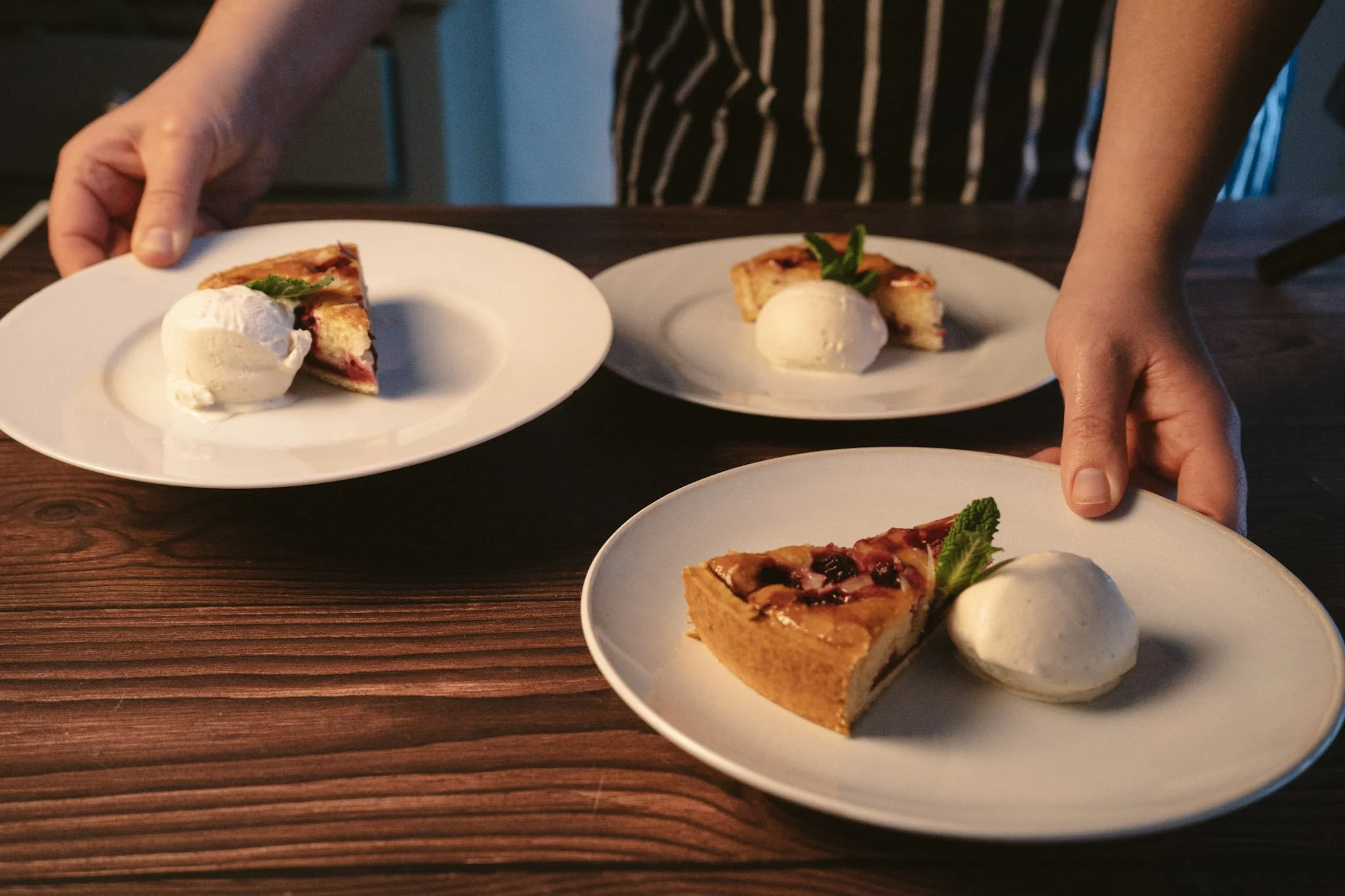 Person serving slices of tart with a scoop of vanilla ice cream on white plates on a wooden table.