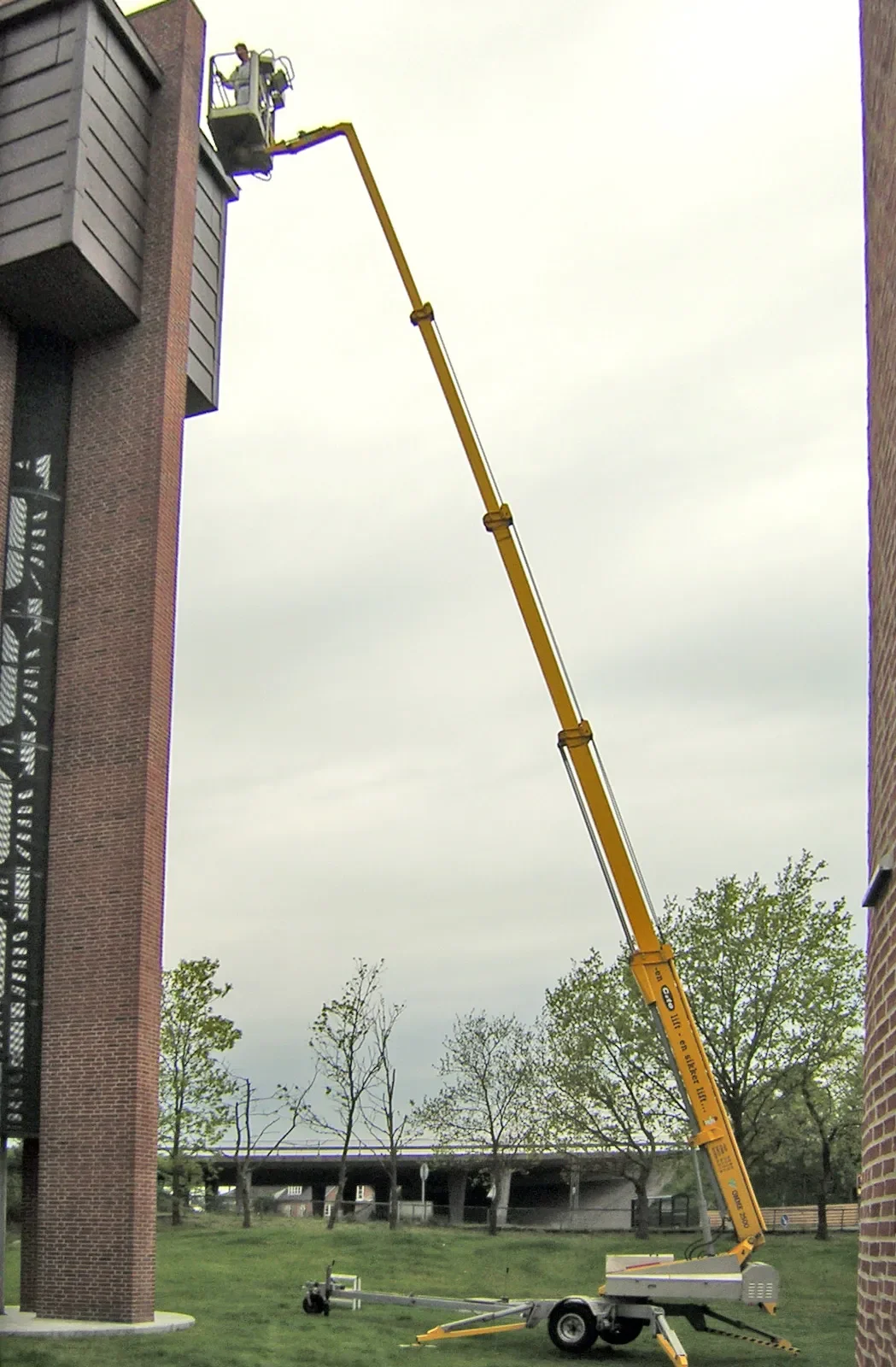 Anhänger-Arbeitsbühne mit sehr langem Teleskopausleger, im Einsatz an einem hohen Gebäude mit Person im Arbeitskorb.