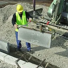 Mechanische Bordsteinversetzzange im Einsatz an einem Hebezeug, geführt von einem Bauarbeiter, zur Miete für das sichere, kraftsparende Heben und exakte Setzen von schweren Bordsteinen im Straßen- und Wegebau.