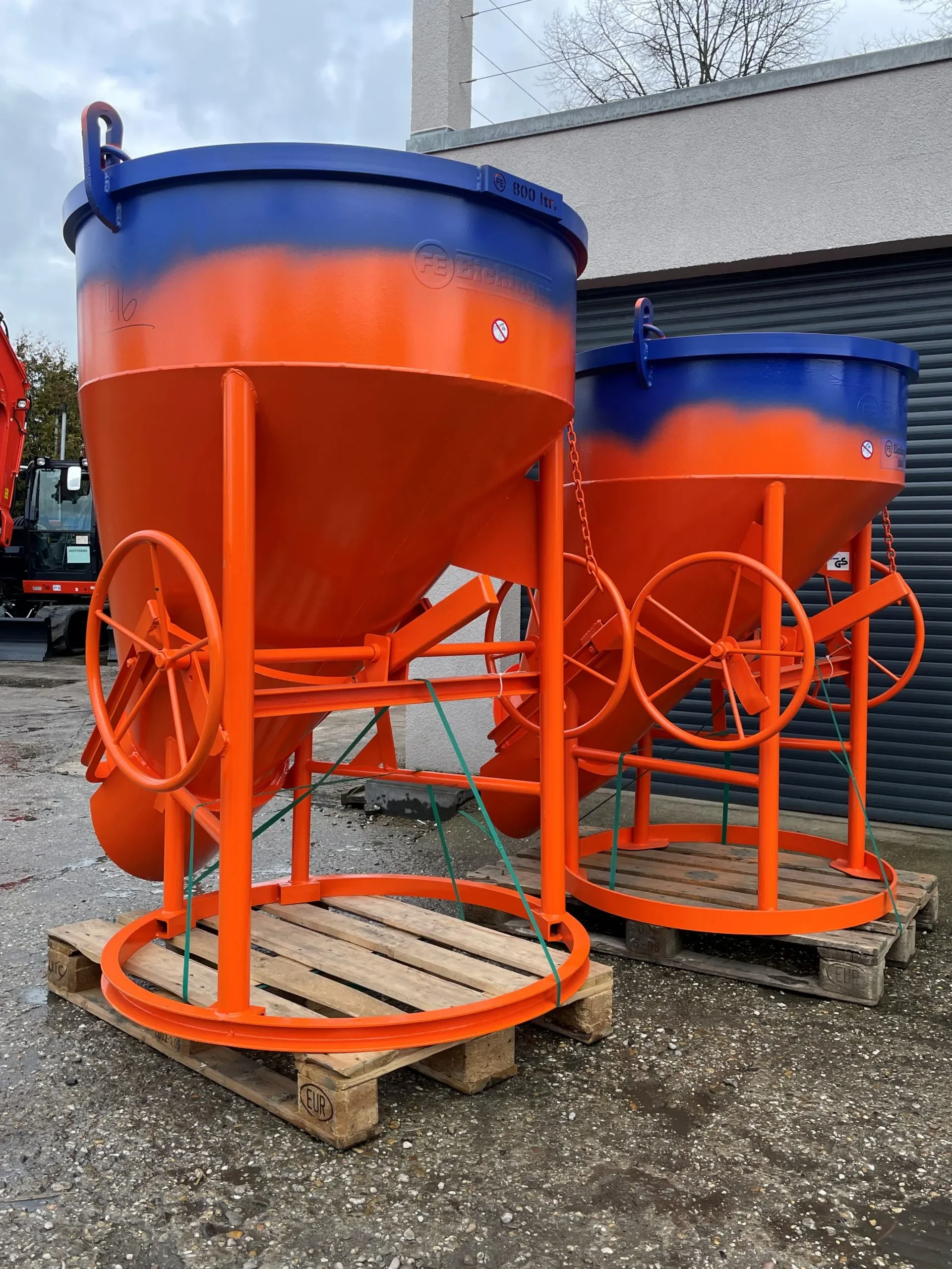 Zwei orange-blaue Betonkübel der Marke Eichinger mit Handrad und Auslaufschurre auf Holzpaletten zur Miete für den sicheren Krantransport und das präzise Gießen von Beton auf Baustellen.