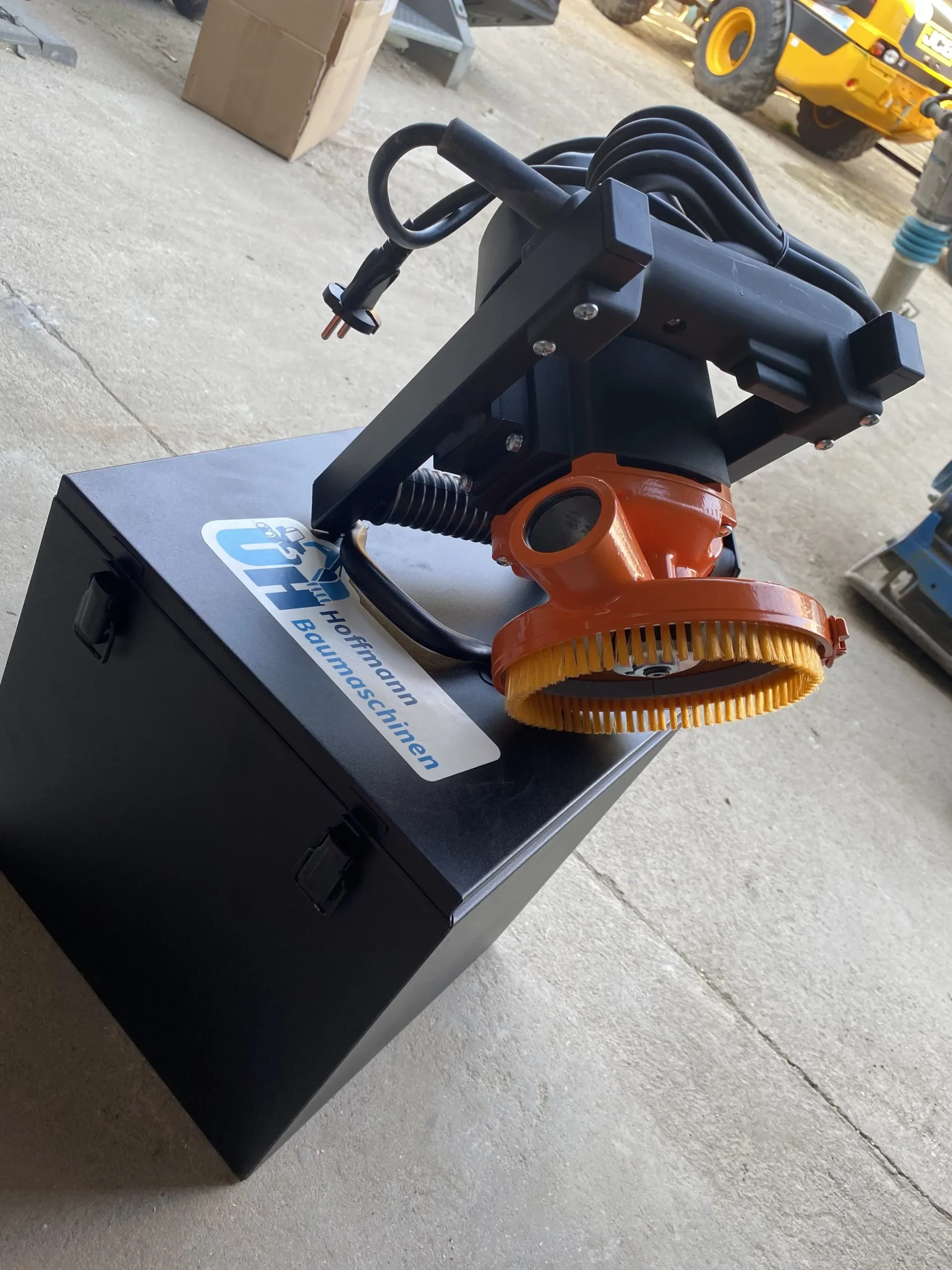 Handgeführter, elektrischer Betonschleifer (Sanierungsschleifer) der Marke Norton mit schwarzem Motorgehäuse, oranger Absaughaube, gelbem Bürstenkranz und robustem Haltegriff, platziert auf einer schwarzen Transportbox von Hoffmann Baumaschinen.