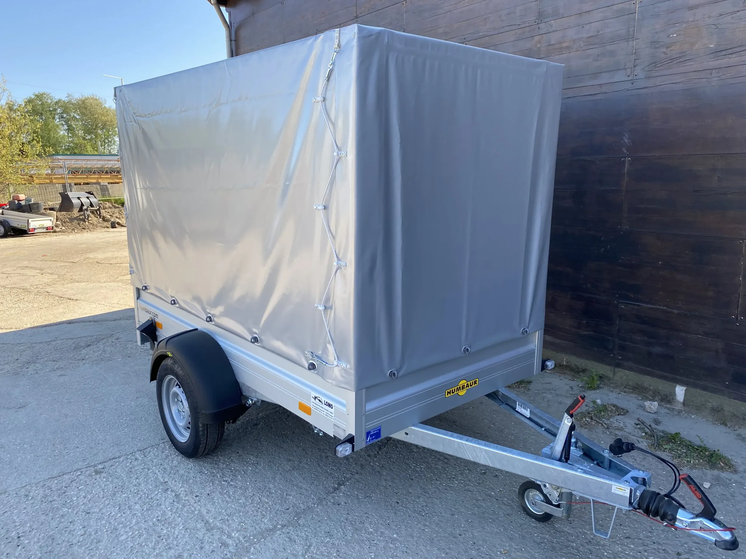 Einachsiger Kastenanhänger mit hoher Plane und Spriegel, abgestellt auf einem Hofgelände.