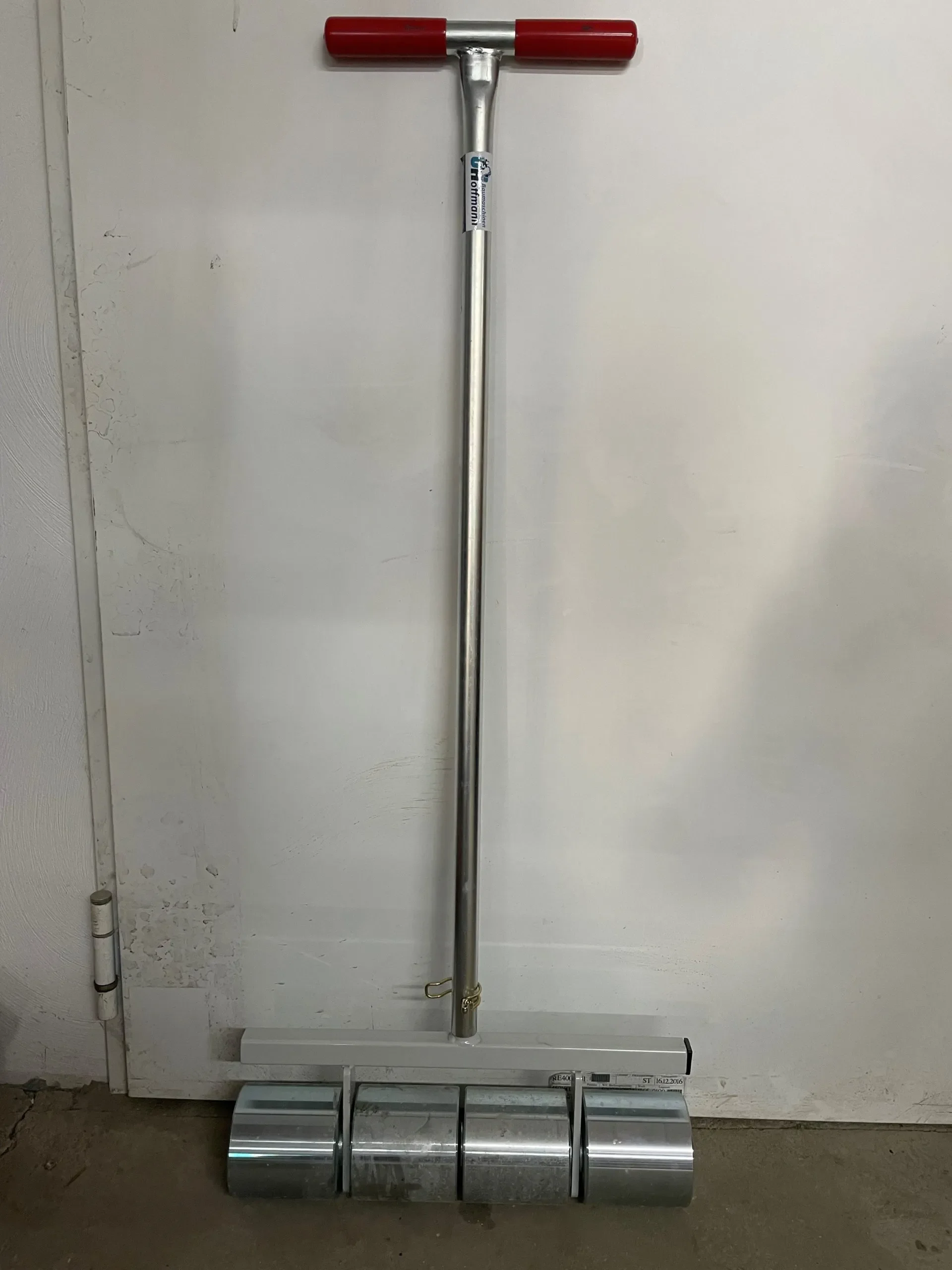 Manuelle, schwere Teppichbodenandrückwalze (Gliederwalze/Anpresswalze) mit langem Metallstiel, rotem T-Griff, vier massiven Metallrollen und Aufkleber von Hoffmann Baumaschinen