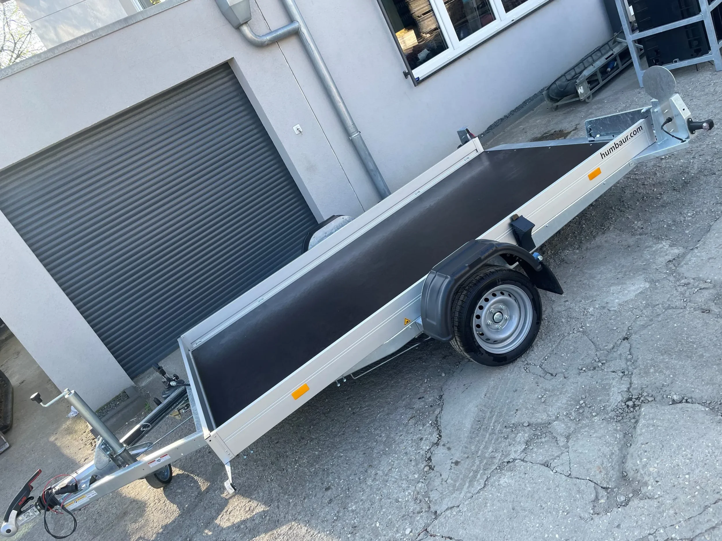 Einachsiger Kastenanhänger mit offener Ladefläche, abgestellt vor einem Gebäude mit Rolltor.