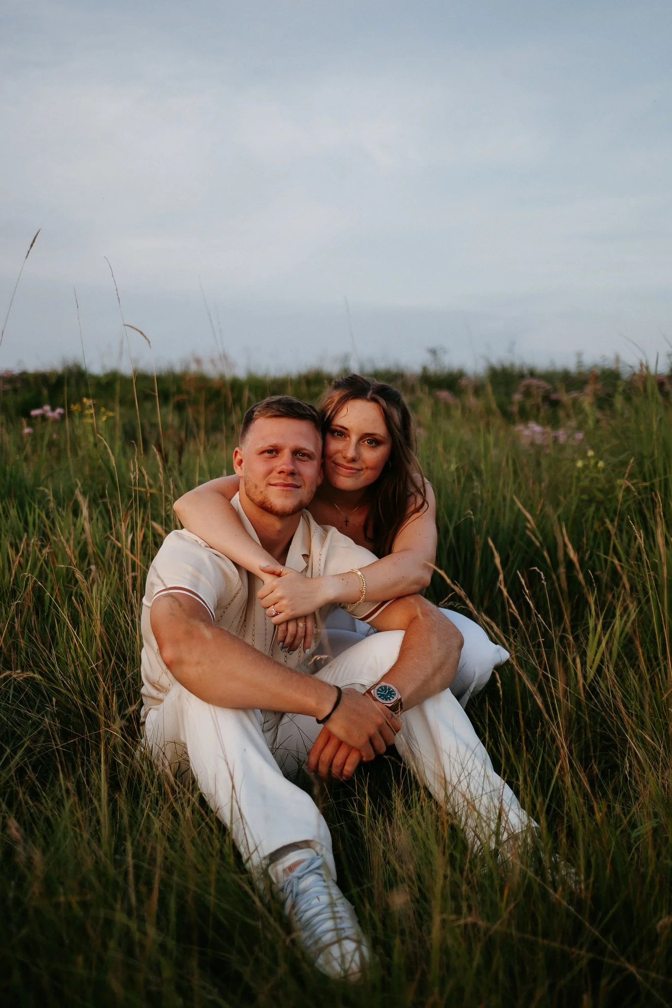 A young couple sitting in tall grass in a field, embracing each other, with a cloudy sky in the background.