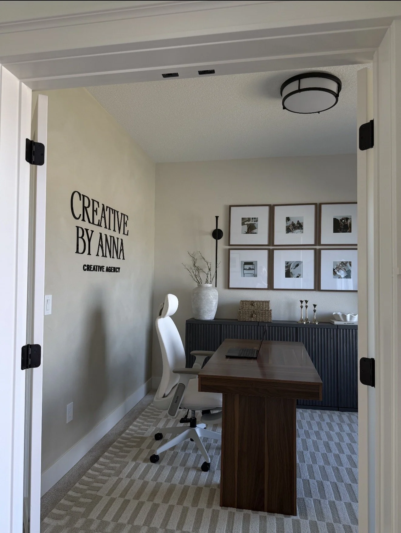 Modern home office with white ergonomic chair, wooden desk, beige and gray patterned rug, and wall decor including framed photos and a statement wall decal that reads 'CREATIVE BY ANNA'.