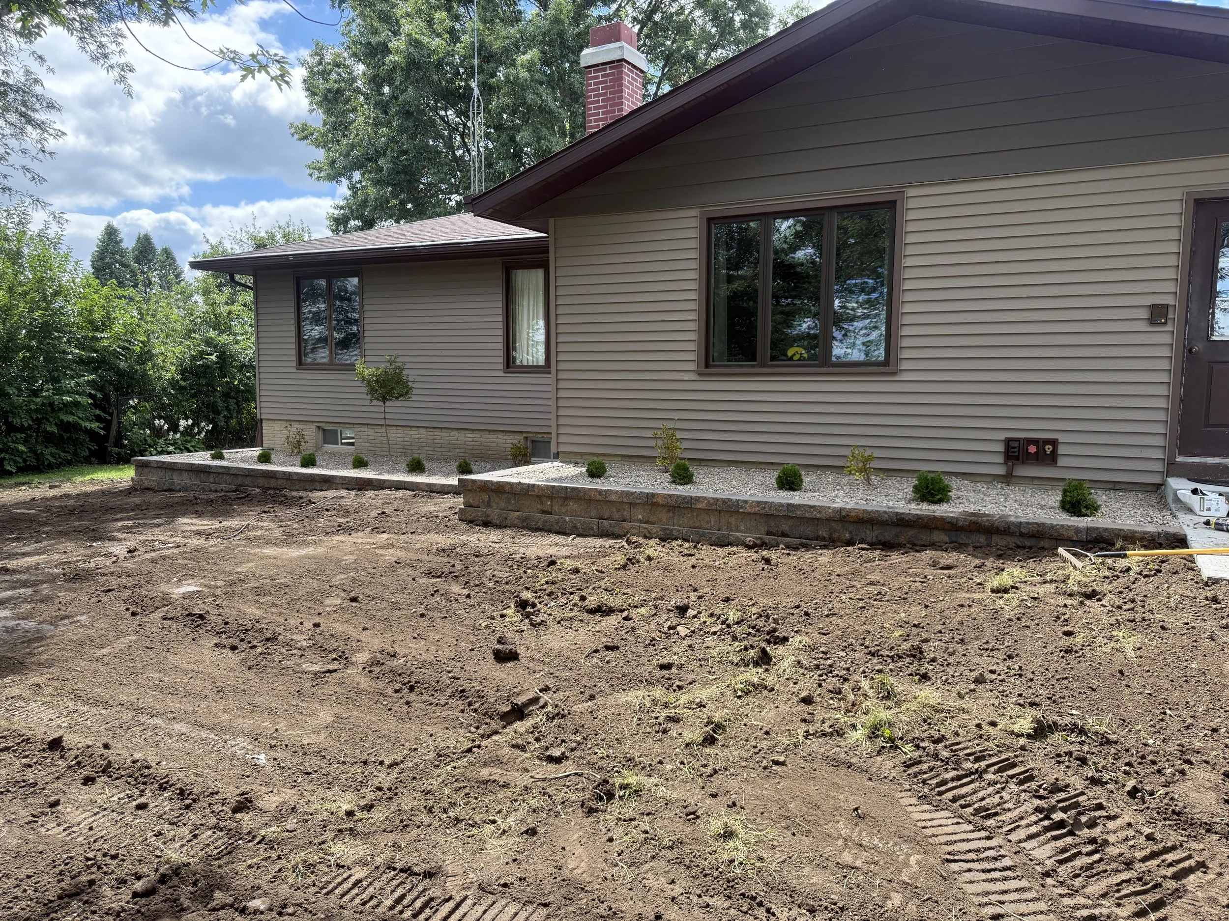 Backyard of a house with newly landscaped garden beds, featuring small green shrubs and a dirt area with tire tracks, and surrounded by trees under a partly cloudy sky.