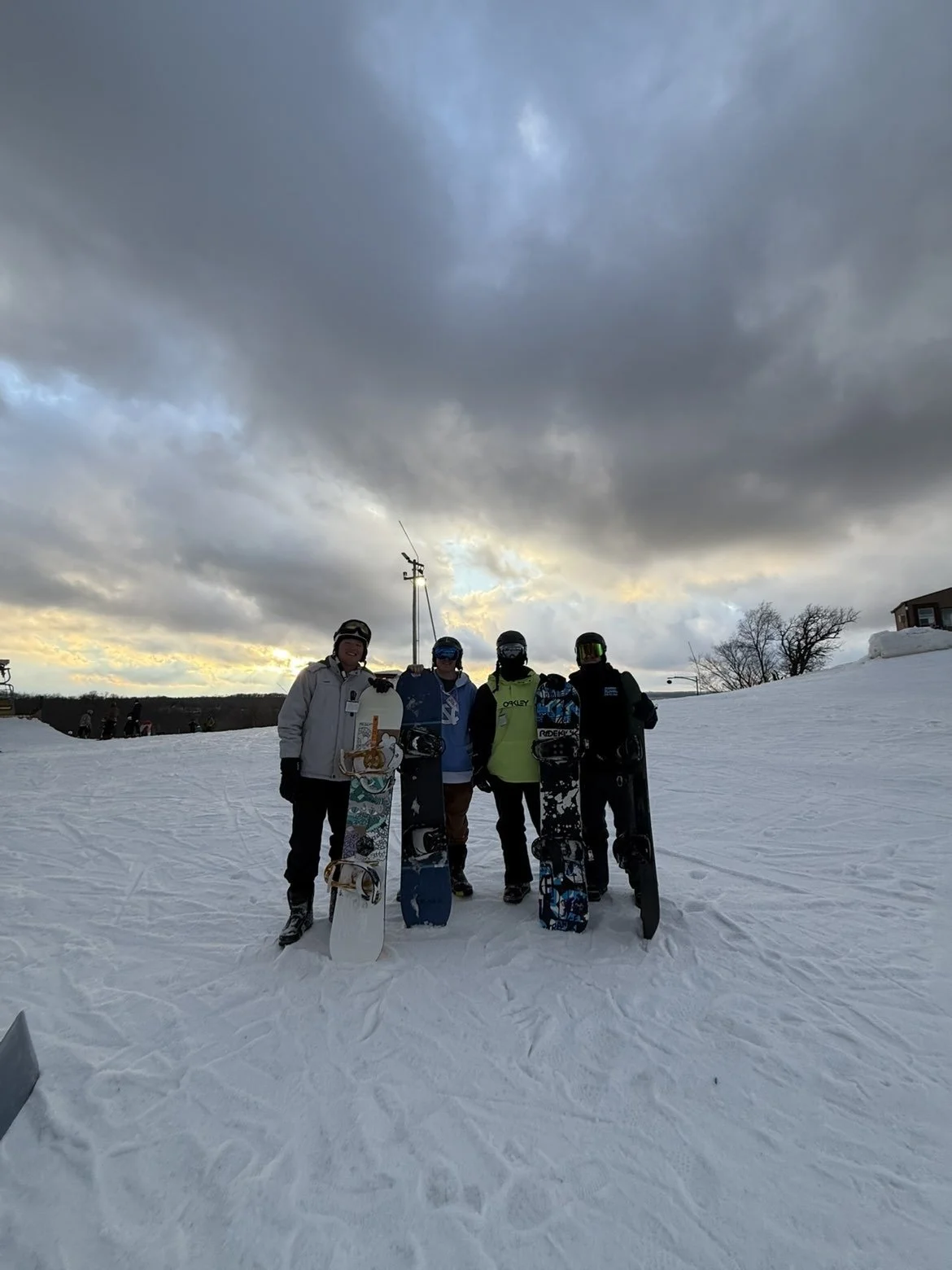 Group of five people in snow gear standing on snow with snowboards, under a cloudy sky at sunset.