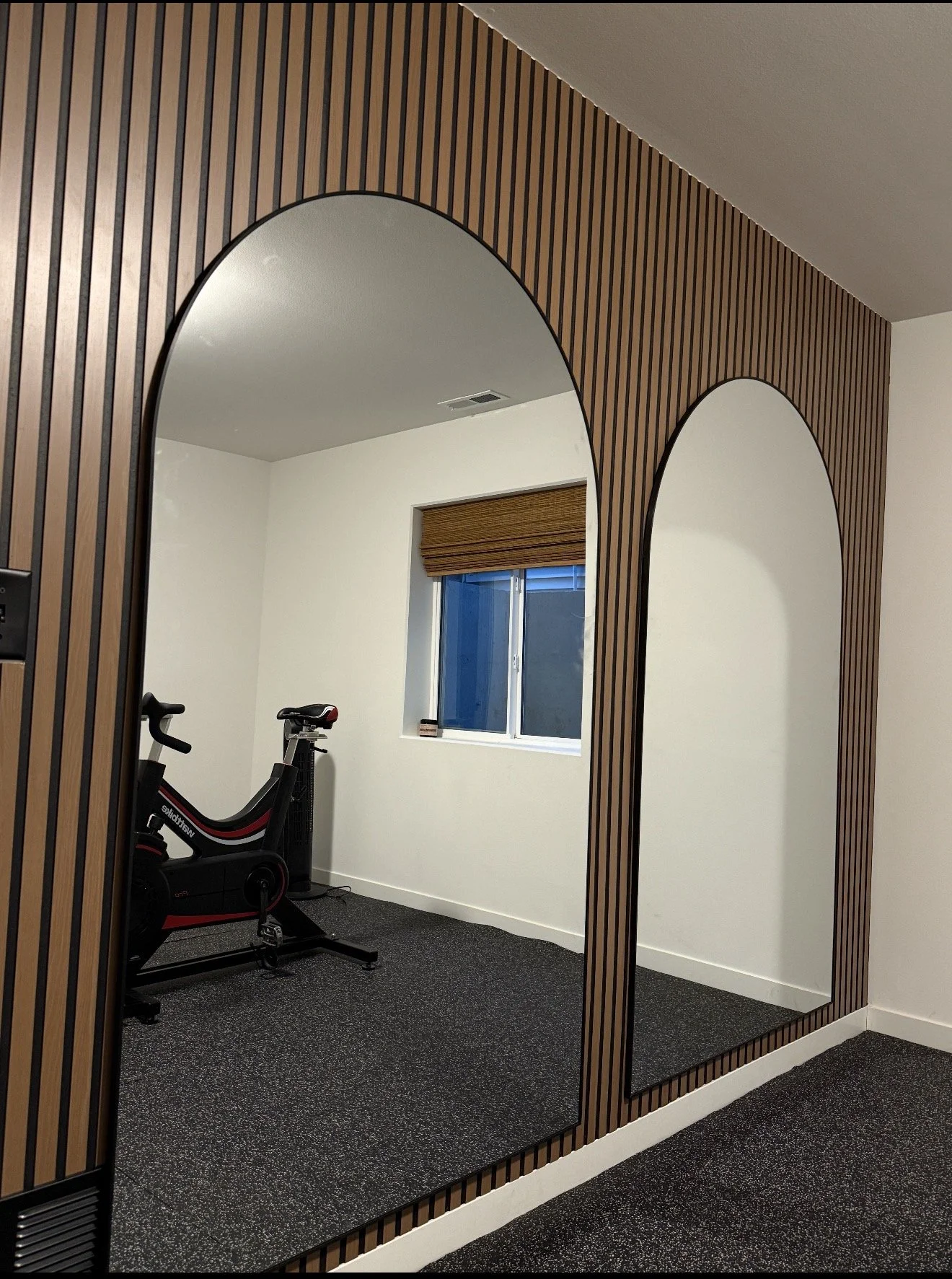 A room with wood-paneled walls and two large mirrors, one arched and the other rectangular, reflecting a window with a brown shade and exercise equipment.