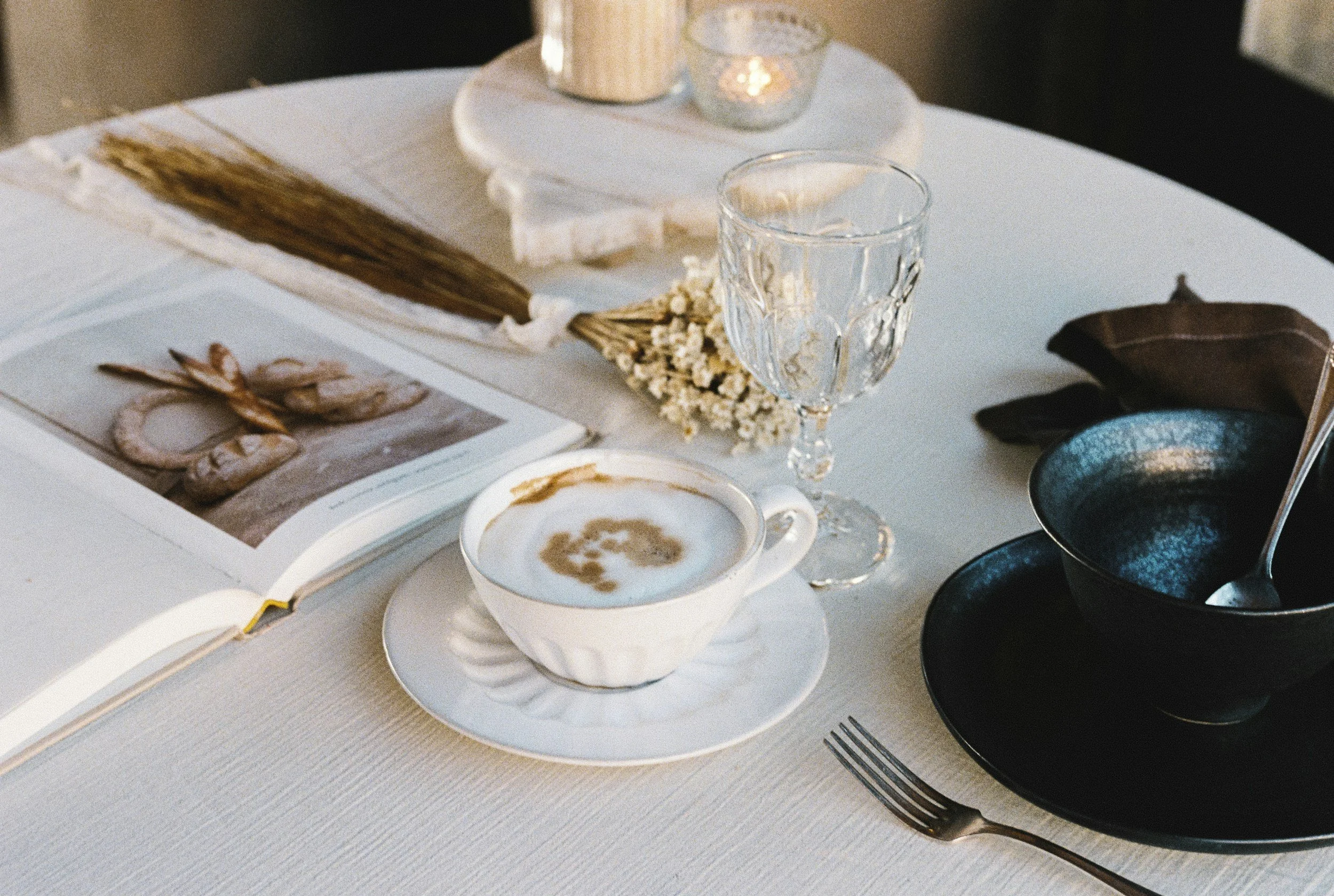 A table with an open book, a cup of coffee, a glass, a black bowl with a spoon, decorative candles, and dried flowers.
