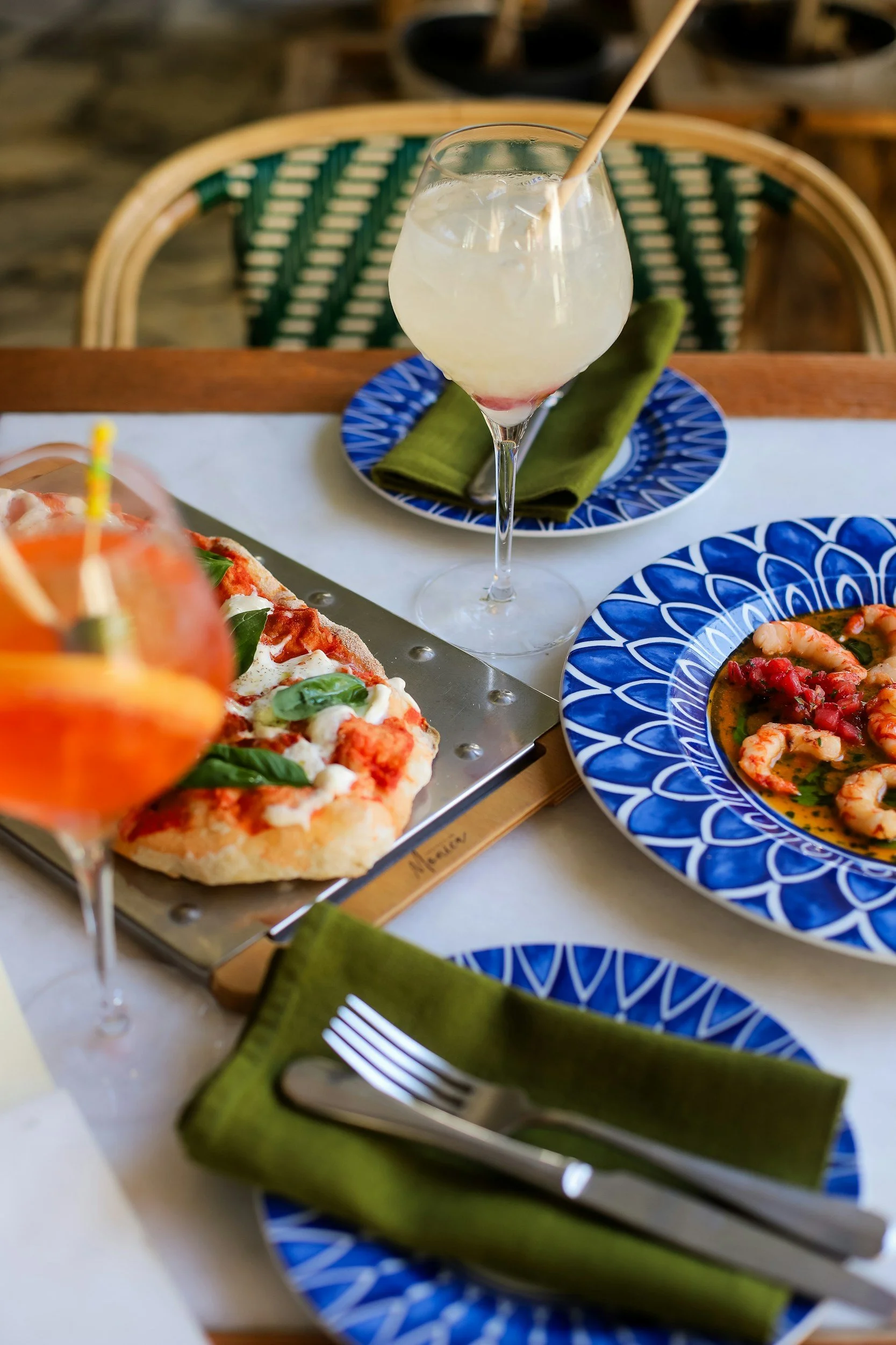 A table set with colorful plates, drinks, and food at a restaurant. There is a slice of pizza with basil and tomato, a seafood dish with shrimp in a sauce, and a cocktail in a glass with a straw. Green napkins and cutlery are also on the table.
