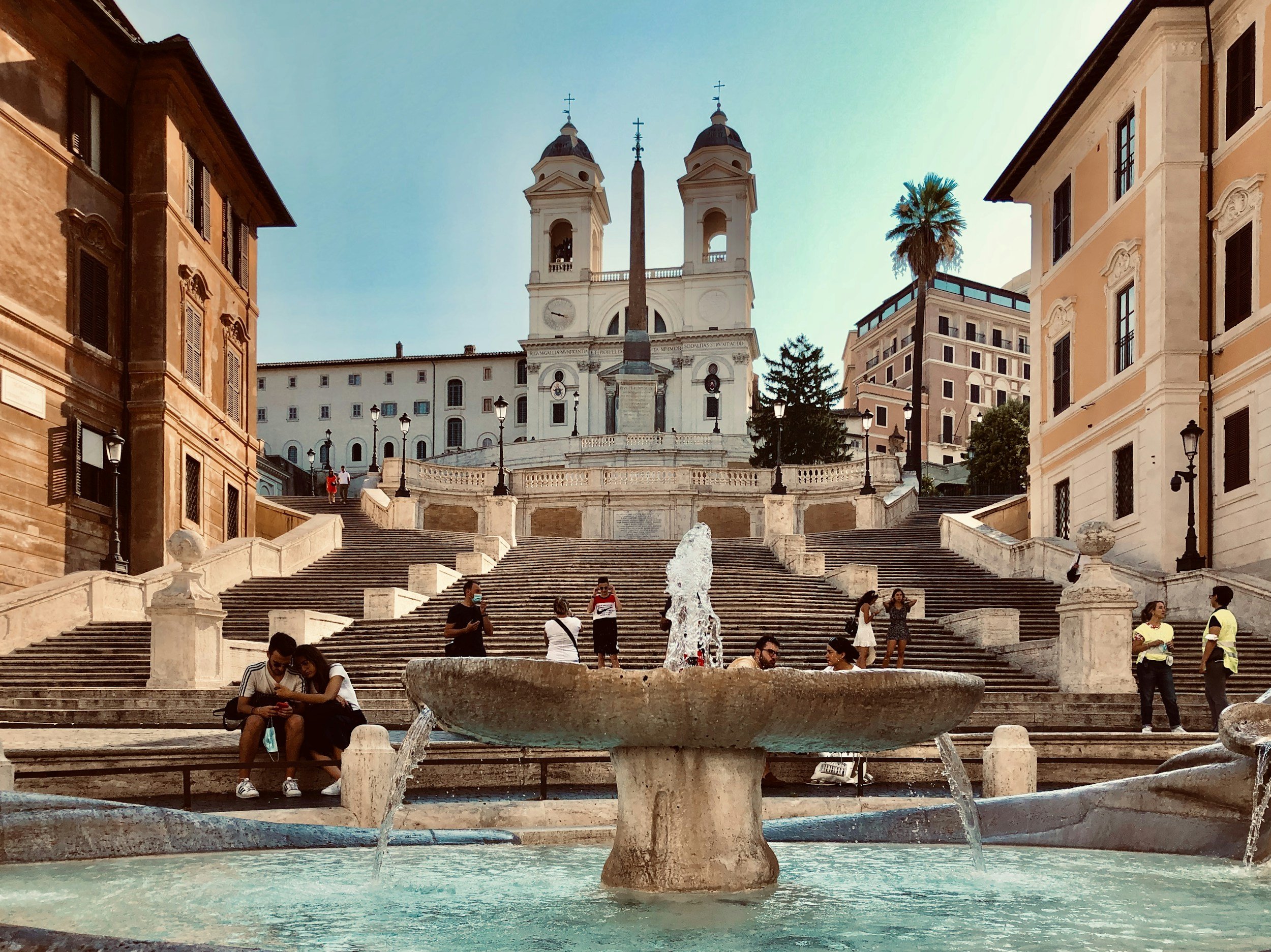 Piazza di Spagna, the Roman most famous square. A historic Roman staircase with a central fountain in front, leading up to a church with twin bell towers, surrounded by colorful buildings, palm trees, and a clear sky.