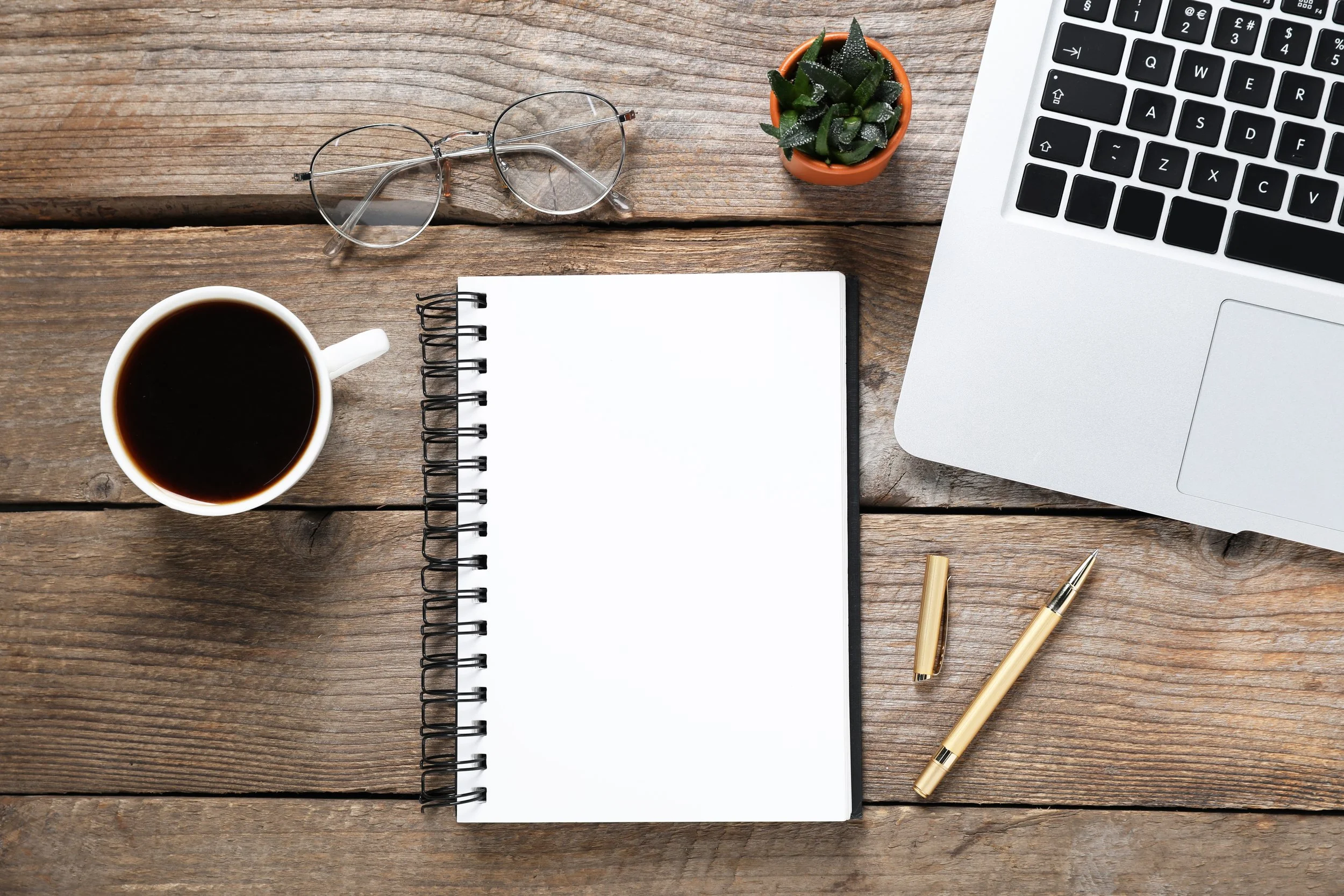 A wooden desk from above with a laptop, open notebook, gold pen, glasses, and a small potted succulent.