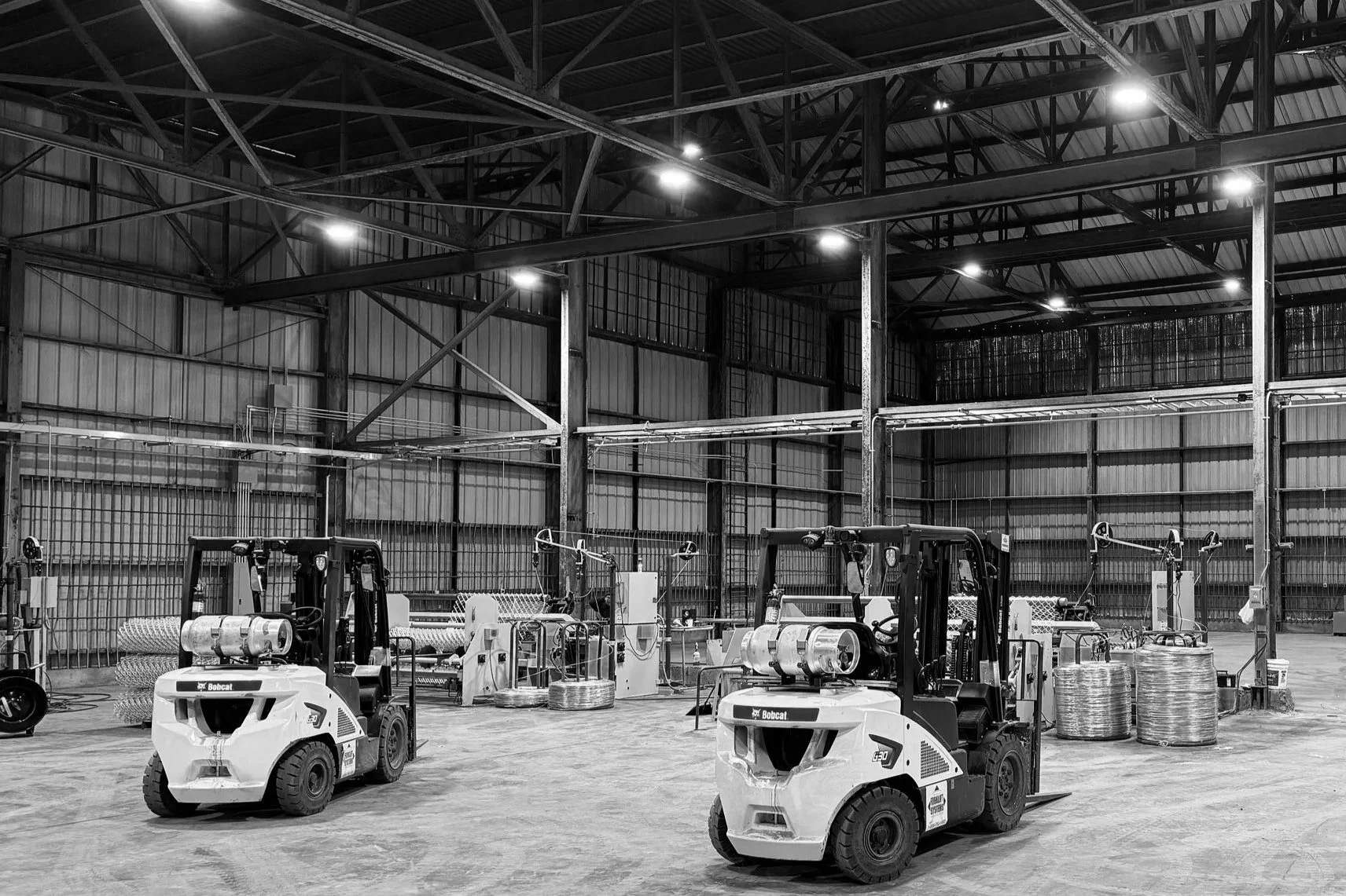 Black and white photo of an industrial warehouse interior with two forklift trucks and large spools of wire or cable on the floor.