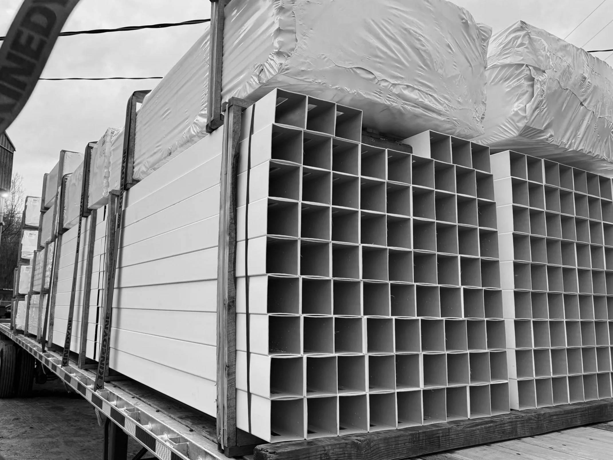 Stacked rectangular white plastic pipes and large wrapped packages on a flatbed truck in a construction site.