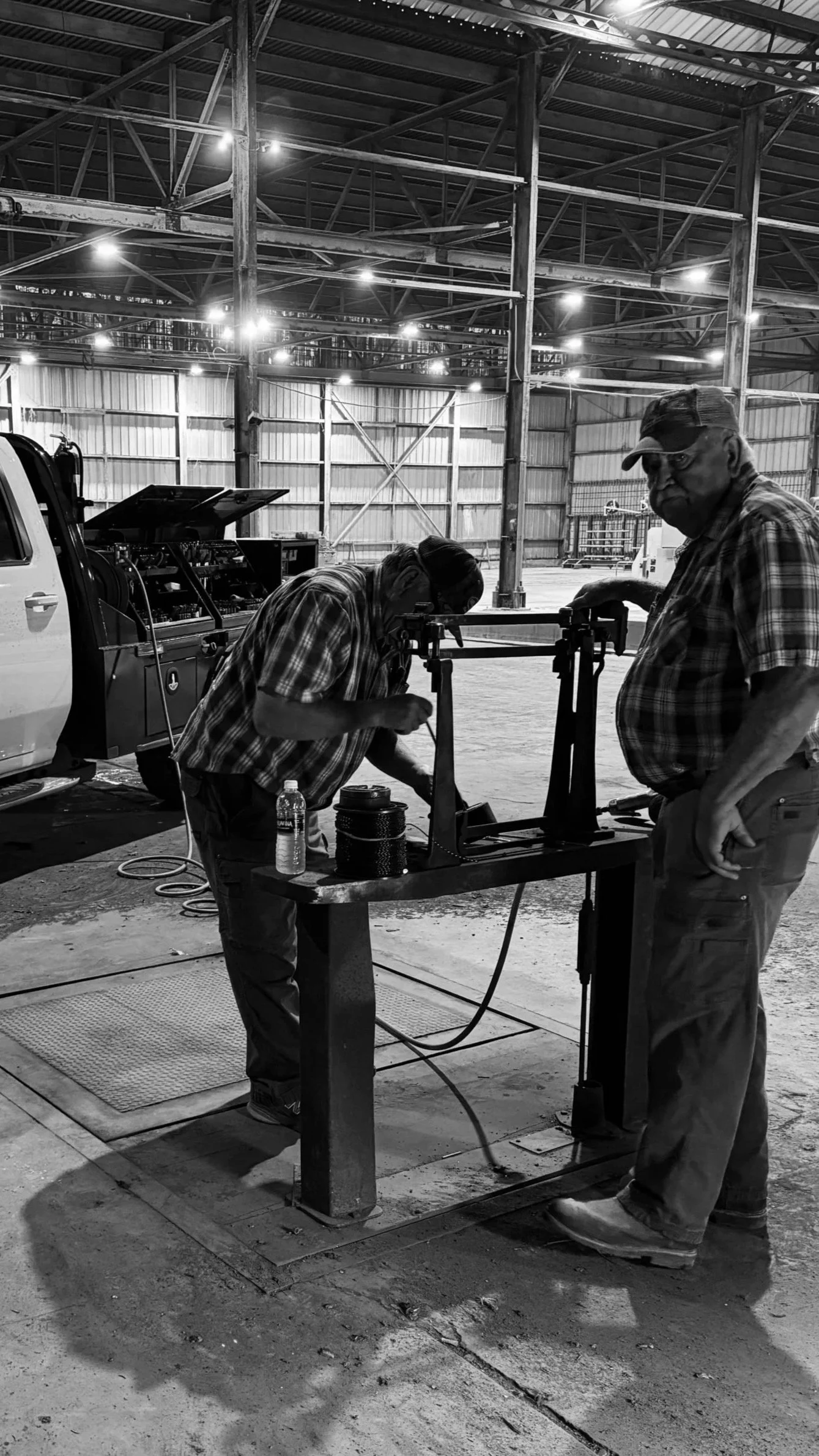 Two men working on a mechanical device in a large indoor warehouse or workshop, with one man examining or adjusting the device while the other observes, both wearing plaid shirts.