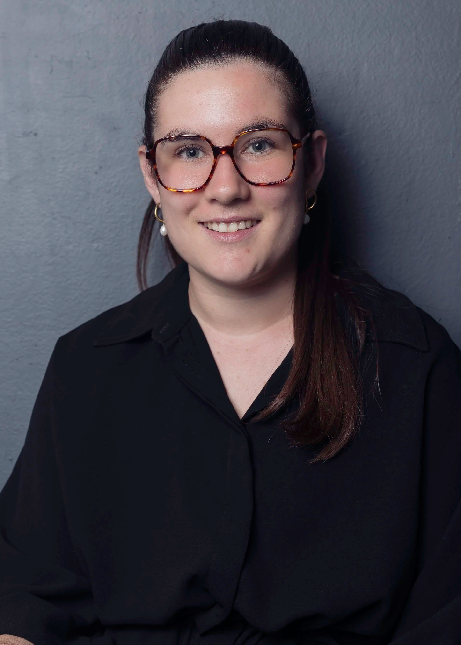 Portrait d'une femme souriante portant des lunettes à monture tortoise, des boucles d'oreilles avec perles, et habillée d'une chemise noire, devant un mur gris.