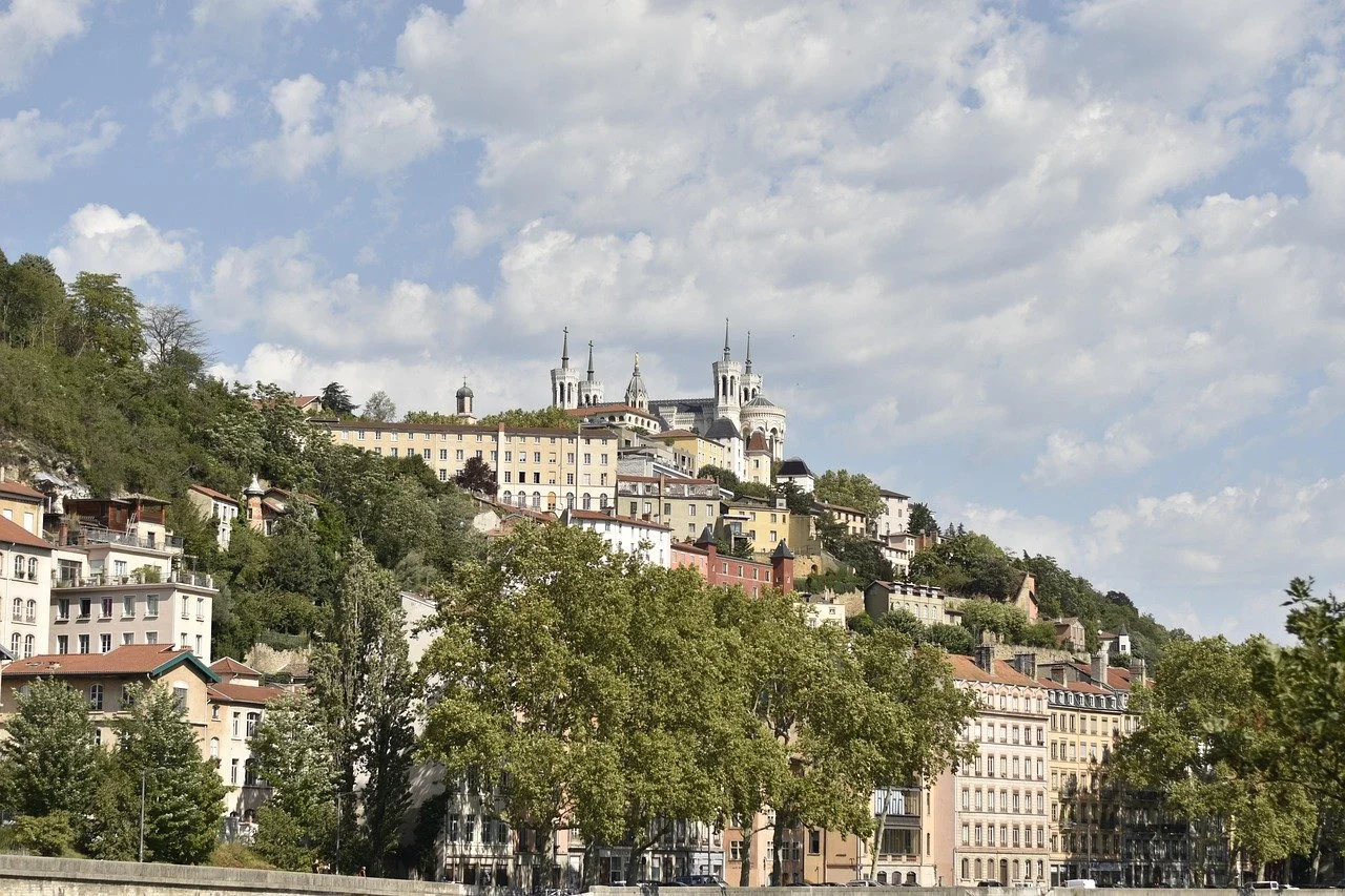 Ville avec maisonnettes colorées et un grand bâtiment blanc en haut de la colline, sous un ciel partiellement nuageux.
