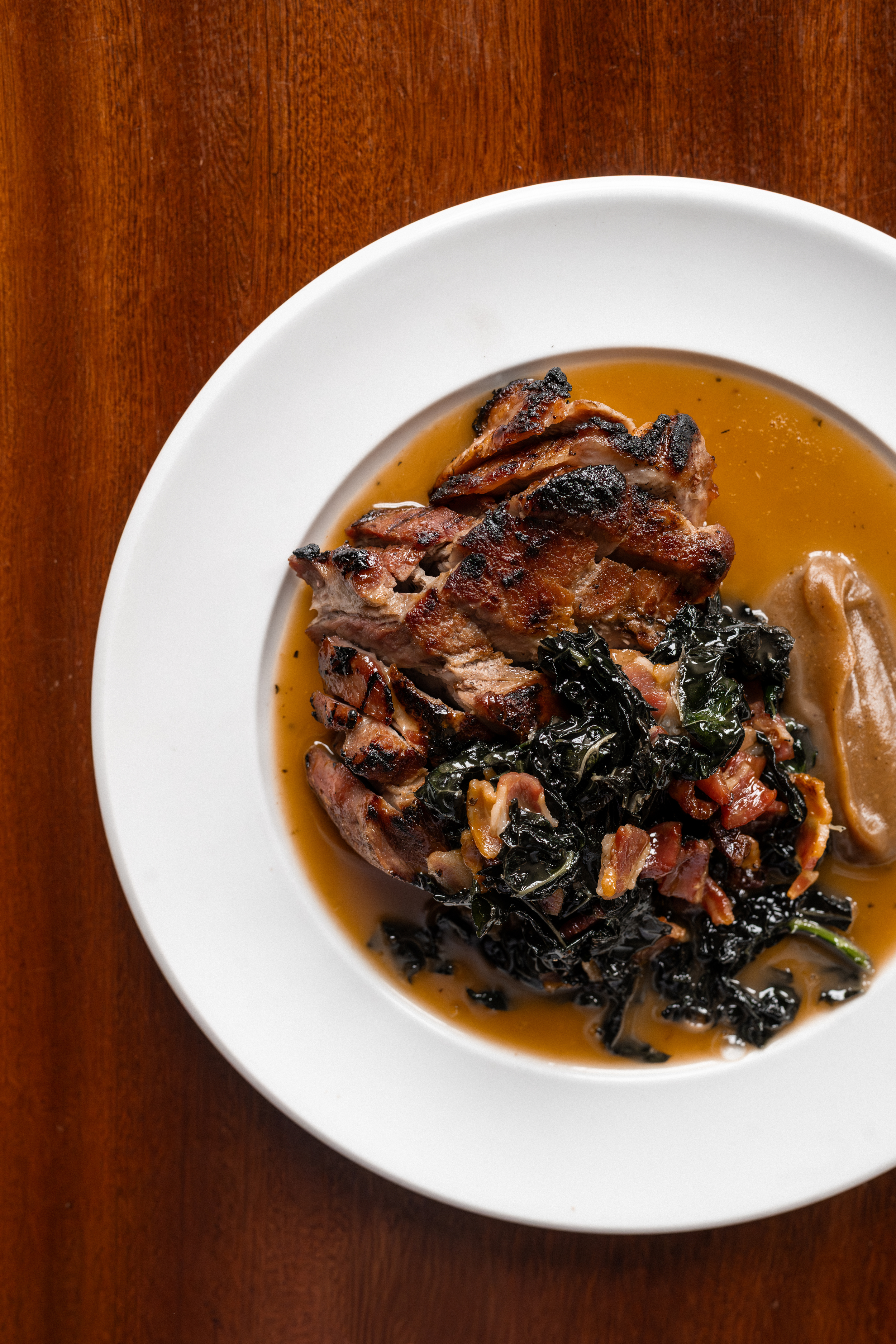 A white plate with grilled pork ribs topped with chopped vegetables and wilted greens, served in a brown gravy on a wooden table.