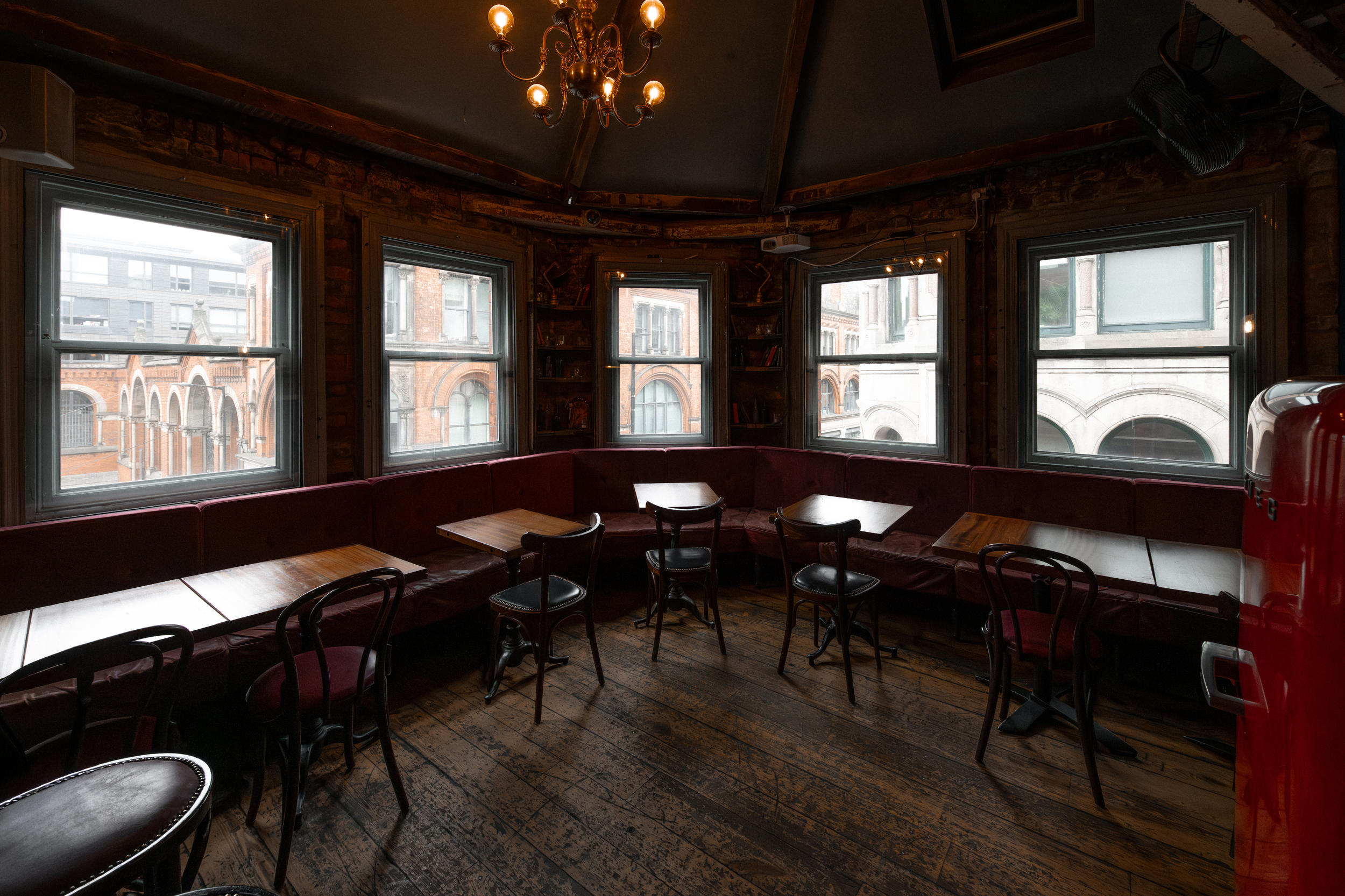Interior of a rustic private room with wooden floors and dark walls, featuring multiple large windows with city buildings visible outside, a red upholstered bench along the wall, small wooden tables, and black chairs.