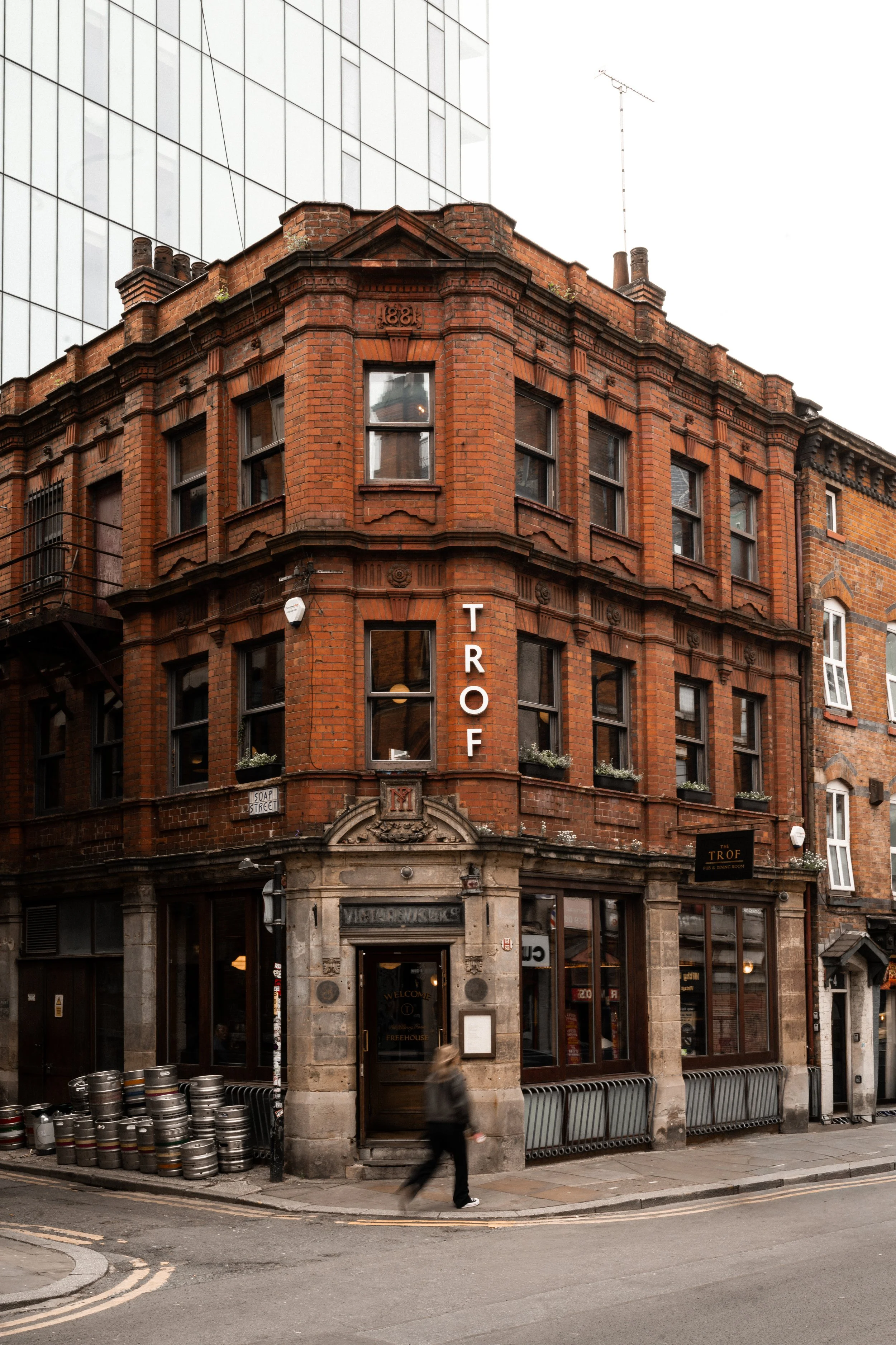A historic red brick building with decorative brickwork and large windows, located on a street corner. The building has a sign for The Trof, a pub and dining room, and is situated next to a modern glass building. The street has barrels stacked outside and a blurred person walking past.