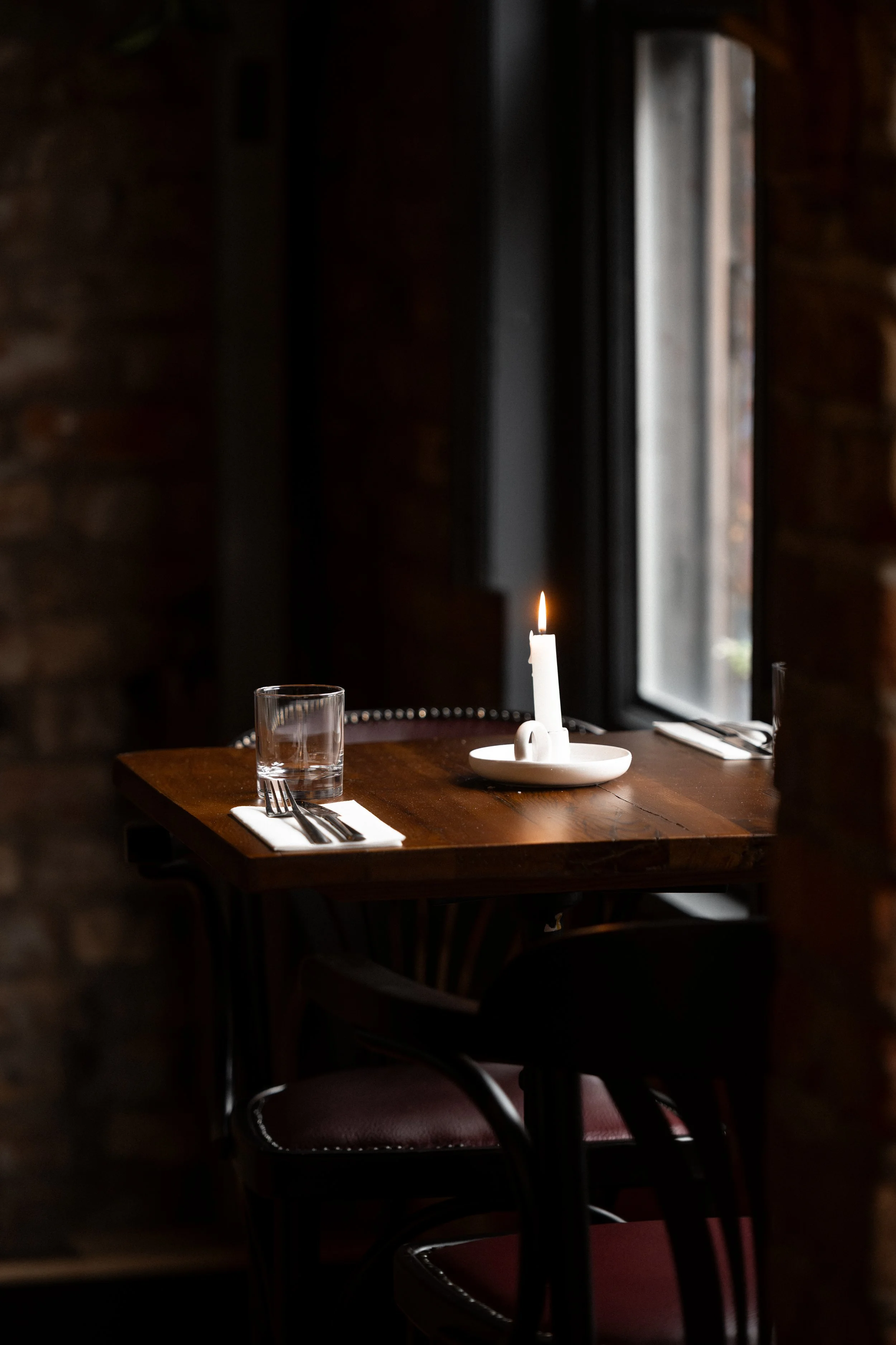 A dimly lit restaurant table set for one, with a white candle in a holder, a glass of water, silverware on a white napkin, and a large window in the background.