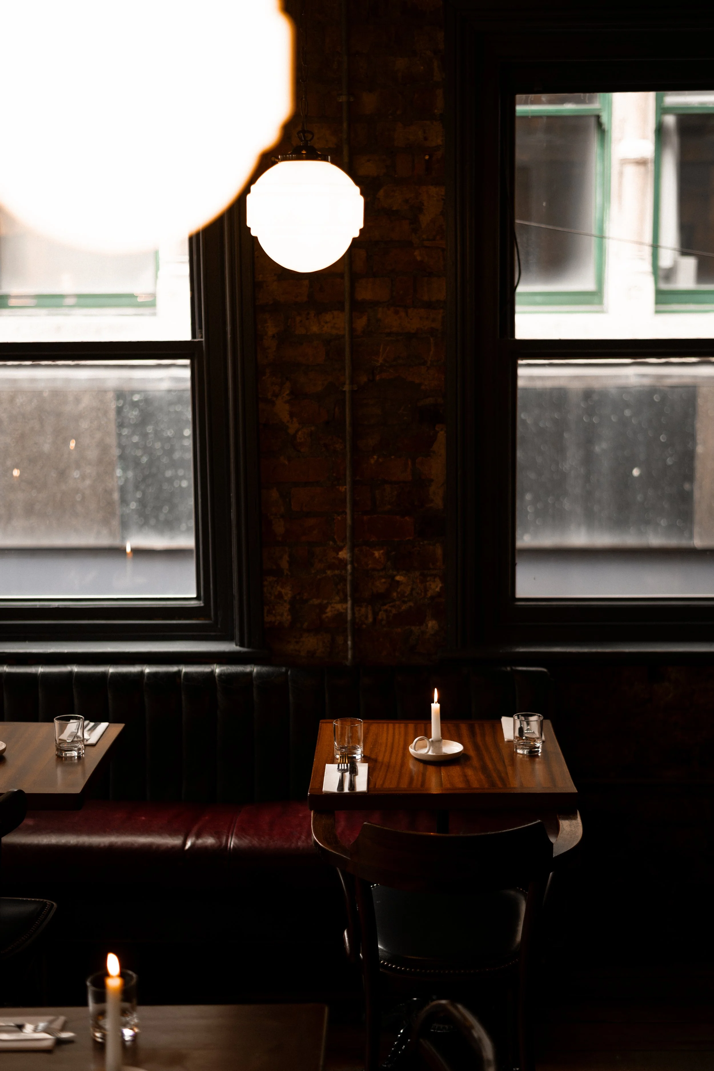 A dinner table with a lit candle, water glasses, and napkins in a restaurant with large windows, brick walls, and hanging lights.
