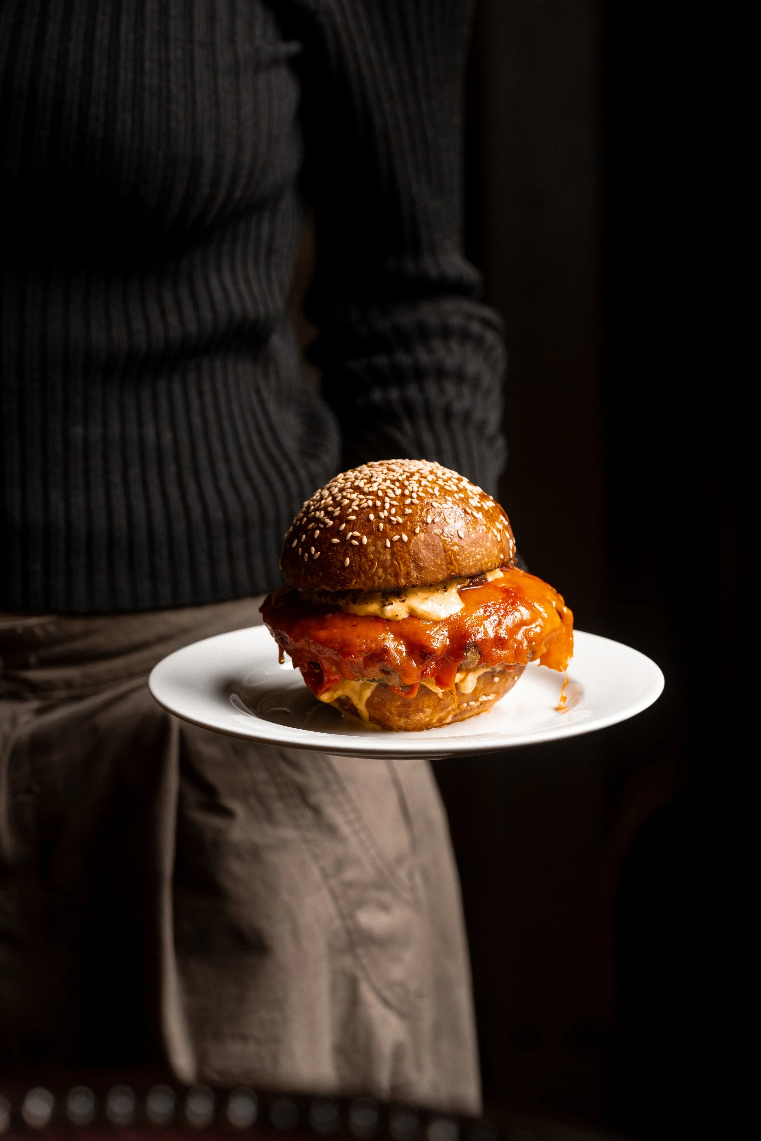 Person holding a white plate with a large burger topped with sesame seed bun, cheese, sauce, and other ingredients, in a dark setting.