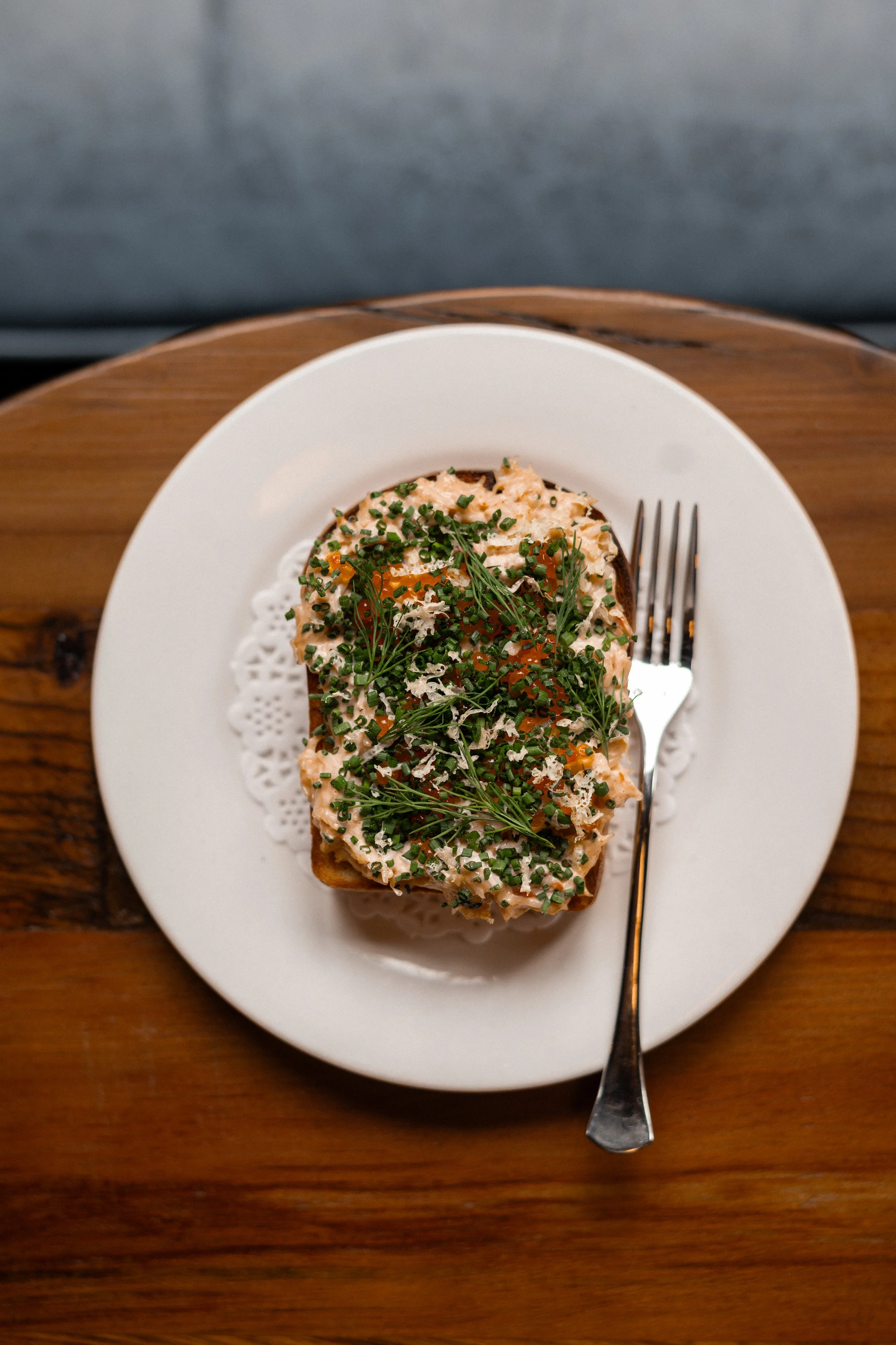 A slice of bread topped with a crab, garnished with chopped chives, and sprigs of fresh herbs, served on a white plate with a fork.