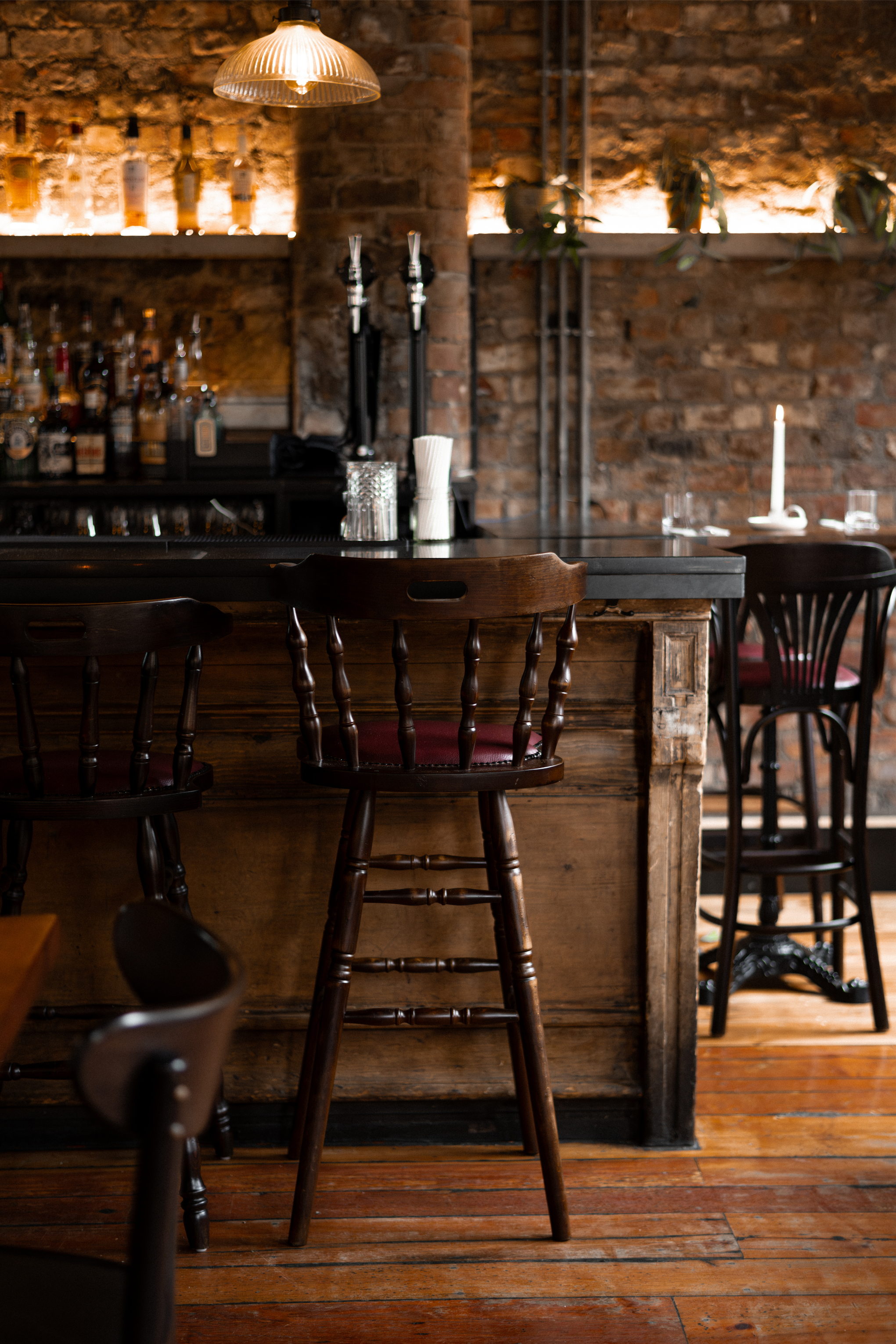 A bar with a brick wall background, featuring a wooden bar counter with bar stools, bottles of alcohol on shelves, a candle, and a hanging lamp.