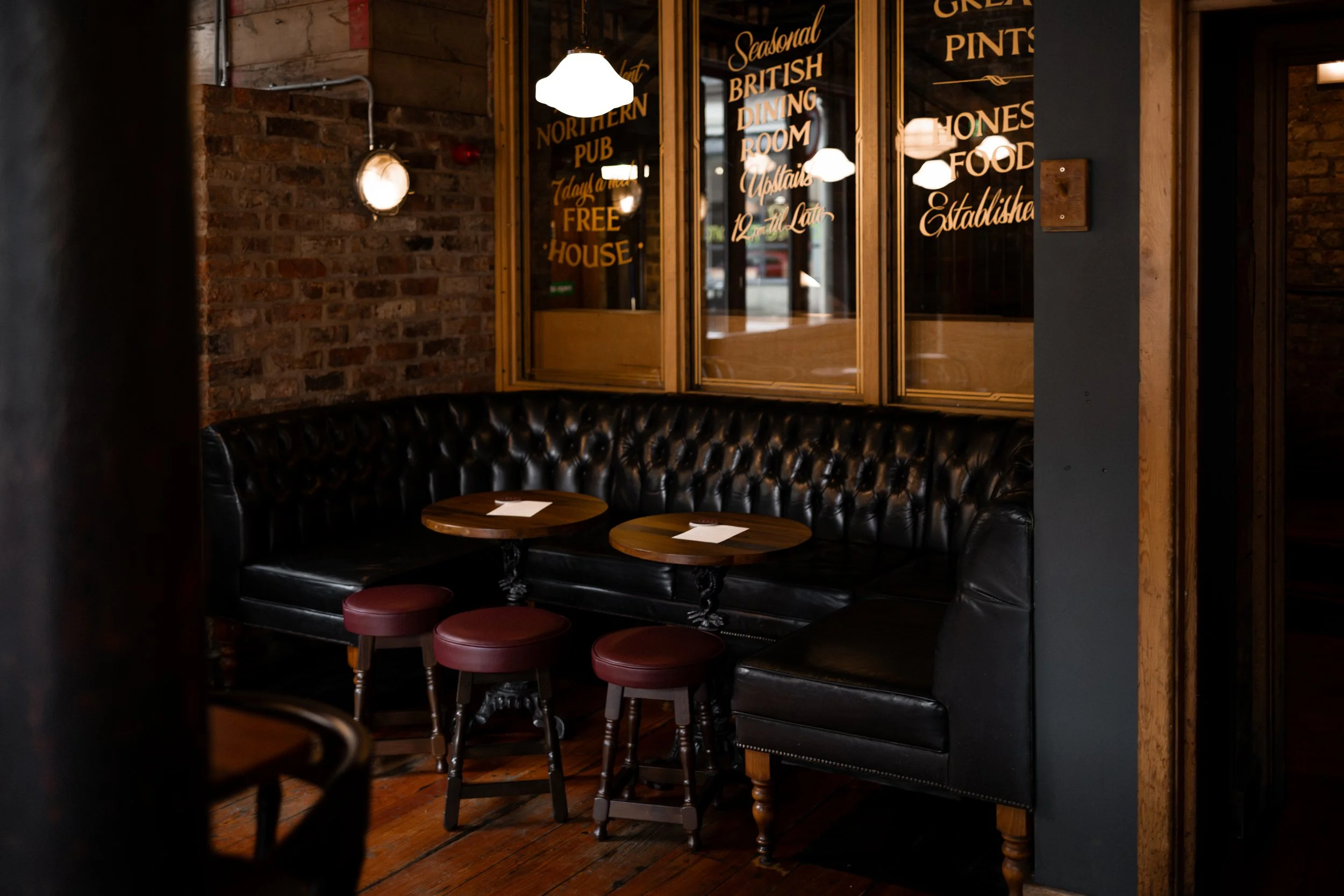 Interior of a cozy pub with brick walls, a black leather banquette, small round tables, and red stools. A window with gold lettering advertising British pub food is visible.
