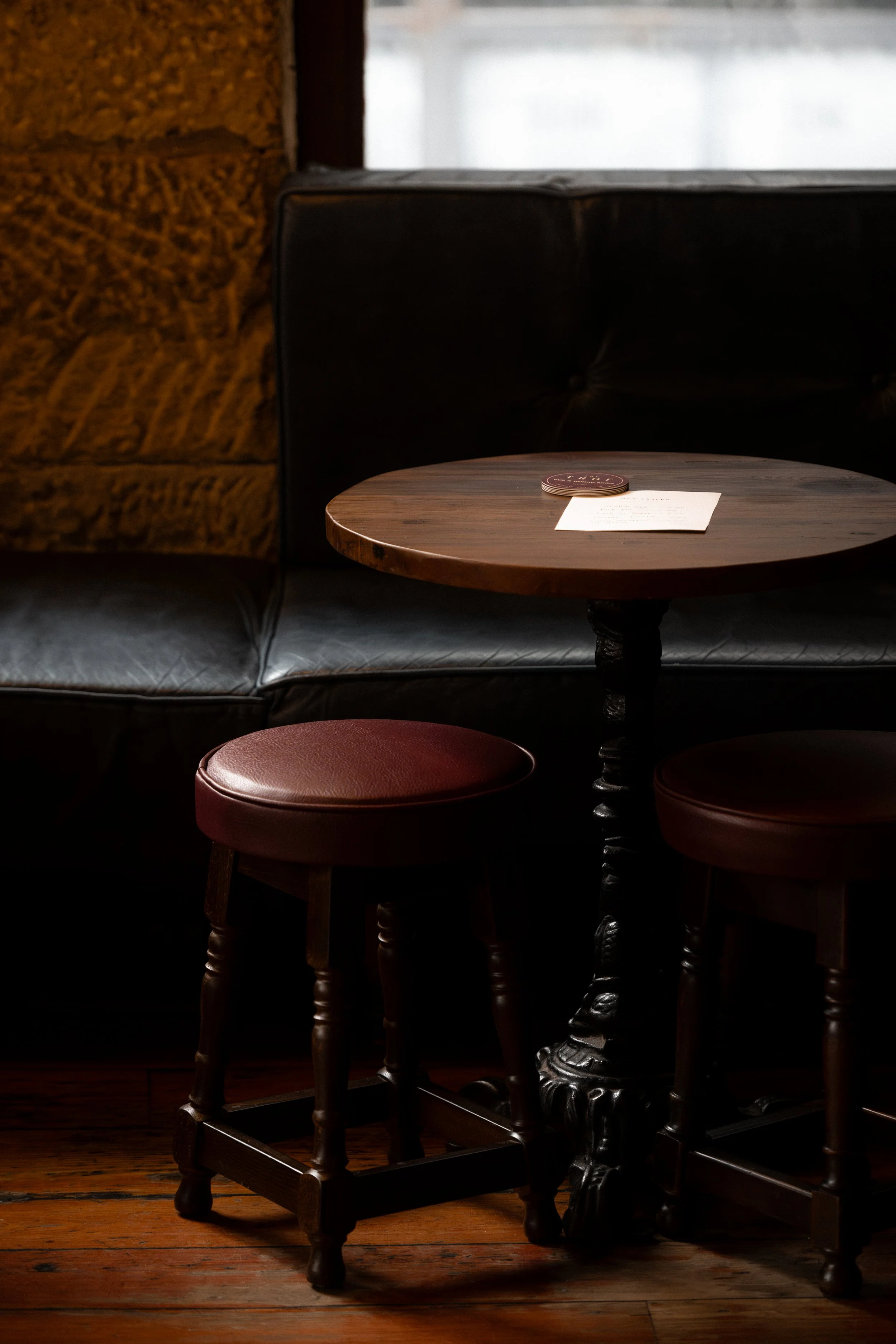 A cozy corner features a round wooden table with a piece of paper and a coaster on it. Surrounding the table are two wooden stools with red cushioned seats. Behind the table, there's a black leather couch with a textured, reddish-brown wall to the left and a window in the background.