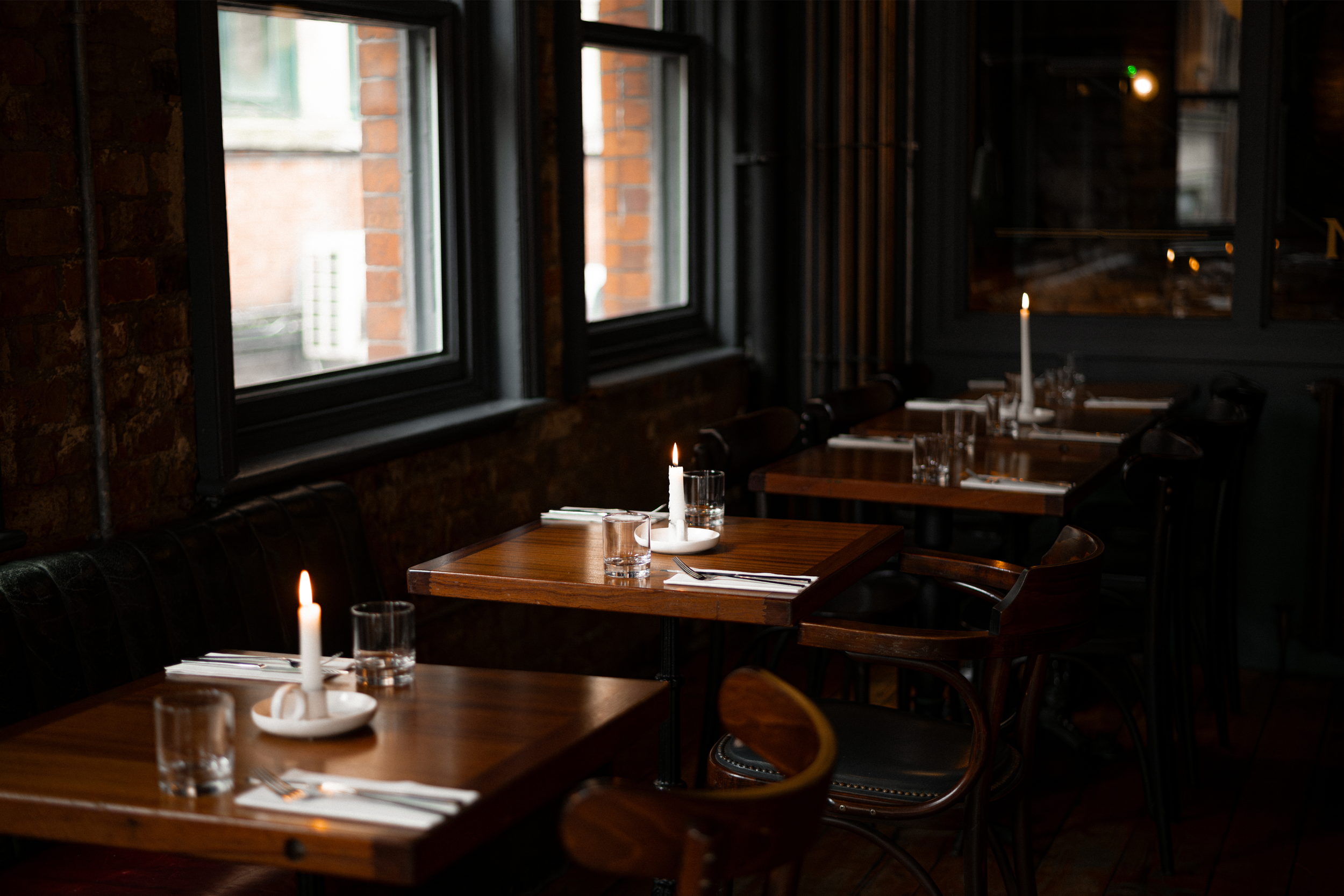 Interior of a dimly lit restaurant with wooden tables, black chairs, and candles as centerpieces, with large windows showing brick buildings outside.