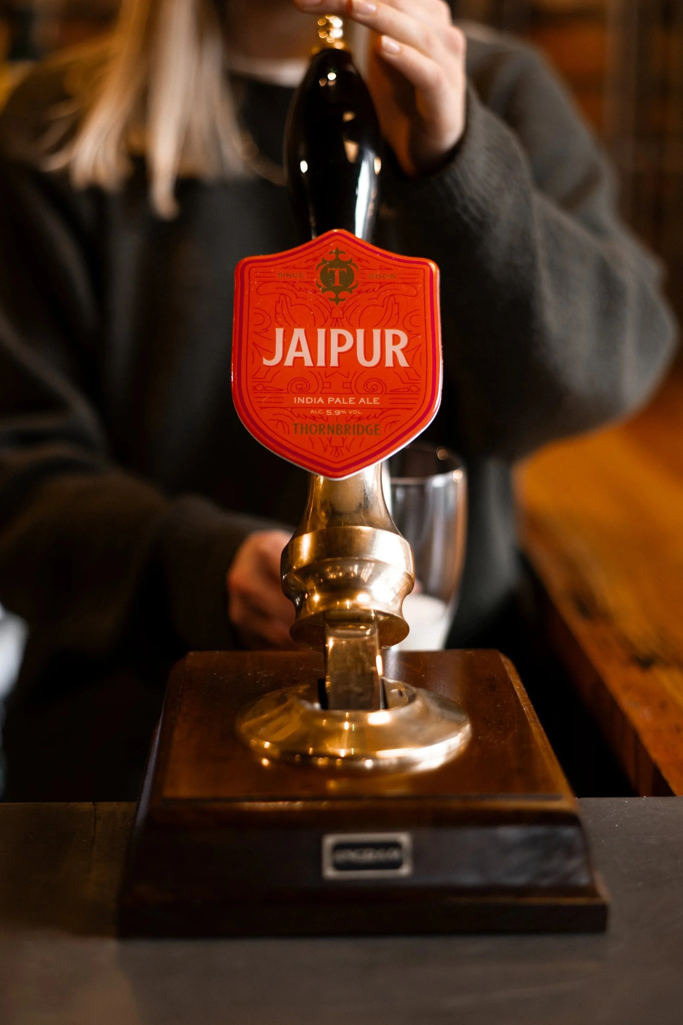 Close-up of a person pulling a beer tap labeled 'Jaipur' on a bar counter, with a blurred background.