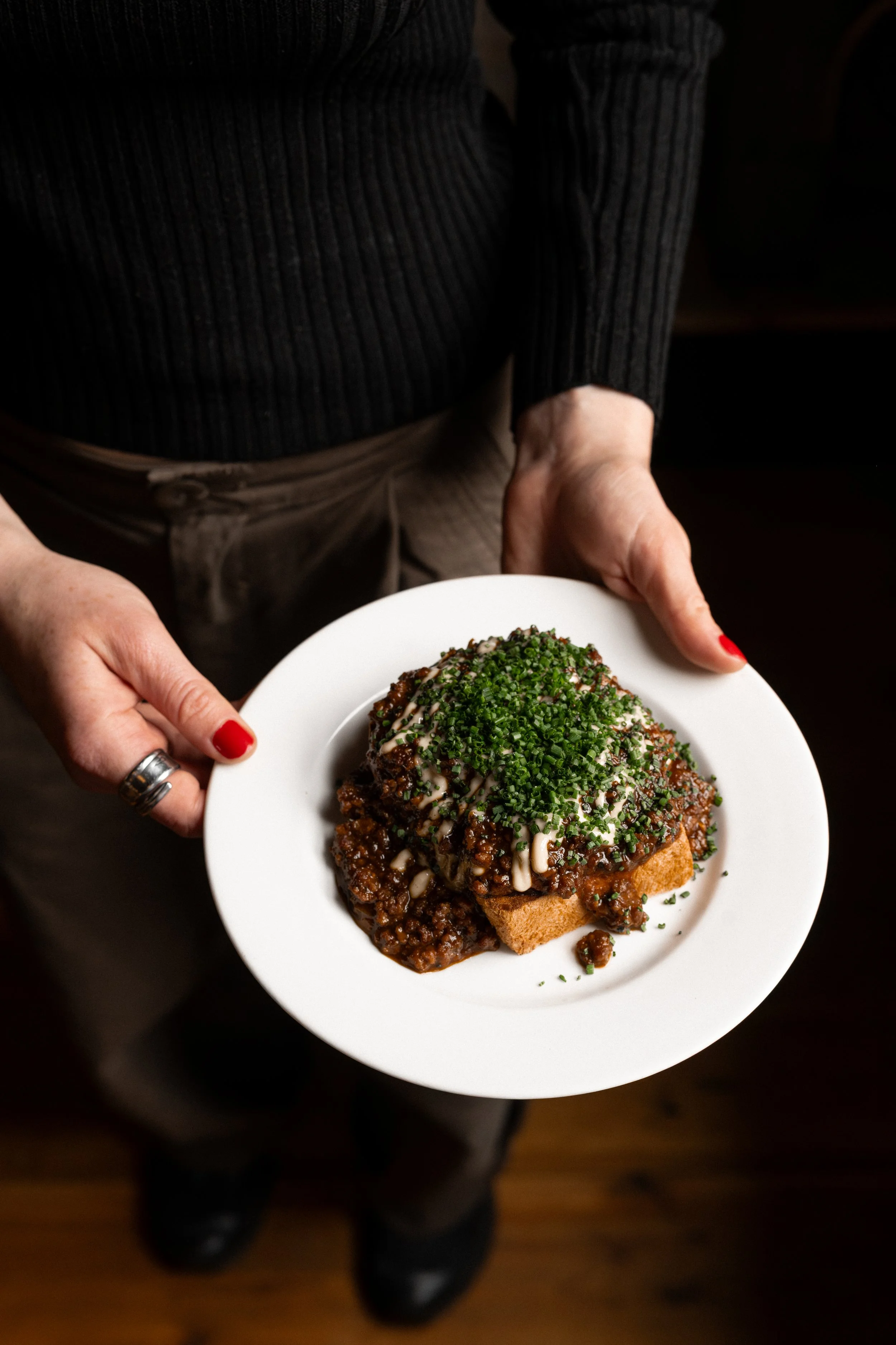 Person holding a white plate with a large serving of topped toast, garnished with chopped chives and drizzle of sauce, wearing a dark, ribbed sweater and brown pants, standing on a wooden floor.