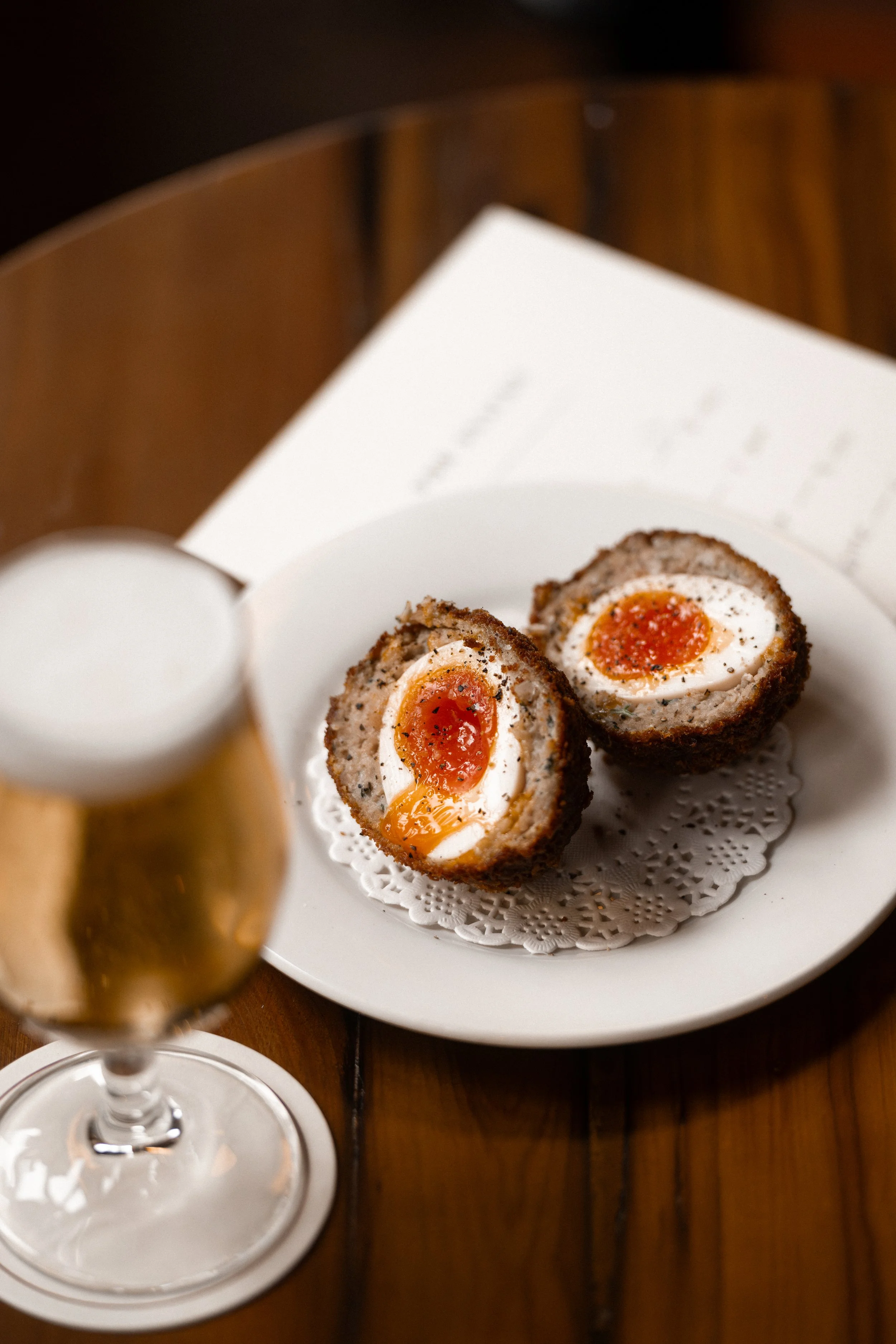 A white plate with two Scotch eggs, with boiled eggs wrapped in seasoned meat and breadcrumbs, served on a doily. In the foreground, a glass of sparkling apple cider on a wooden table.