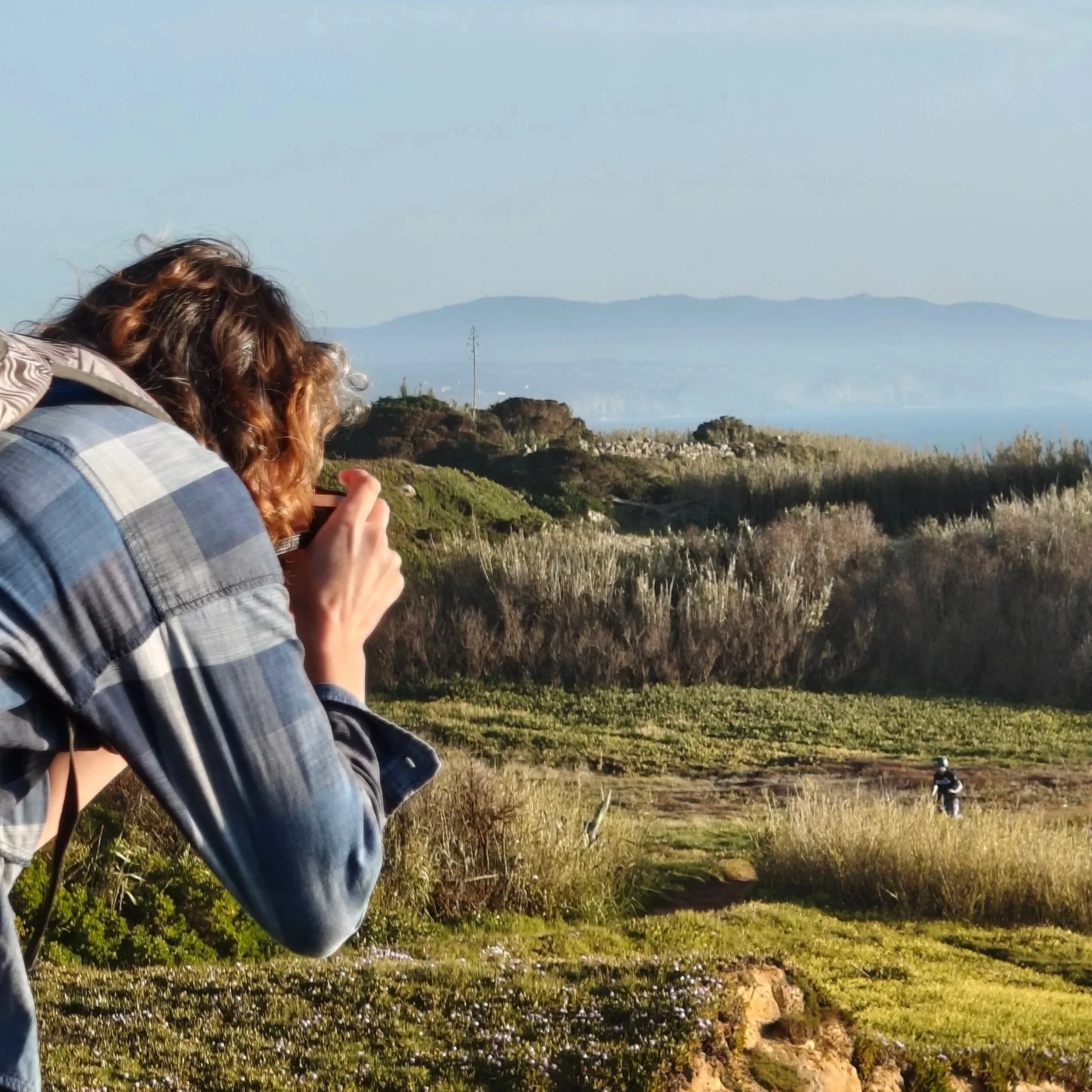 Woman taking a photo in nature with a man in the midground and the ocean and mountain in the background