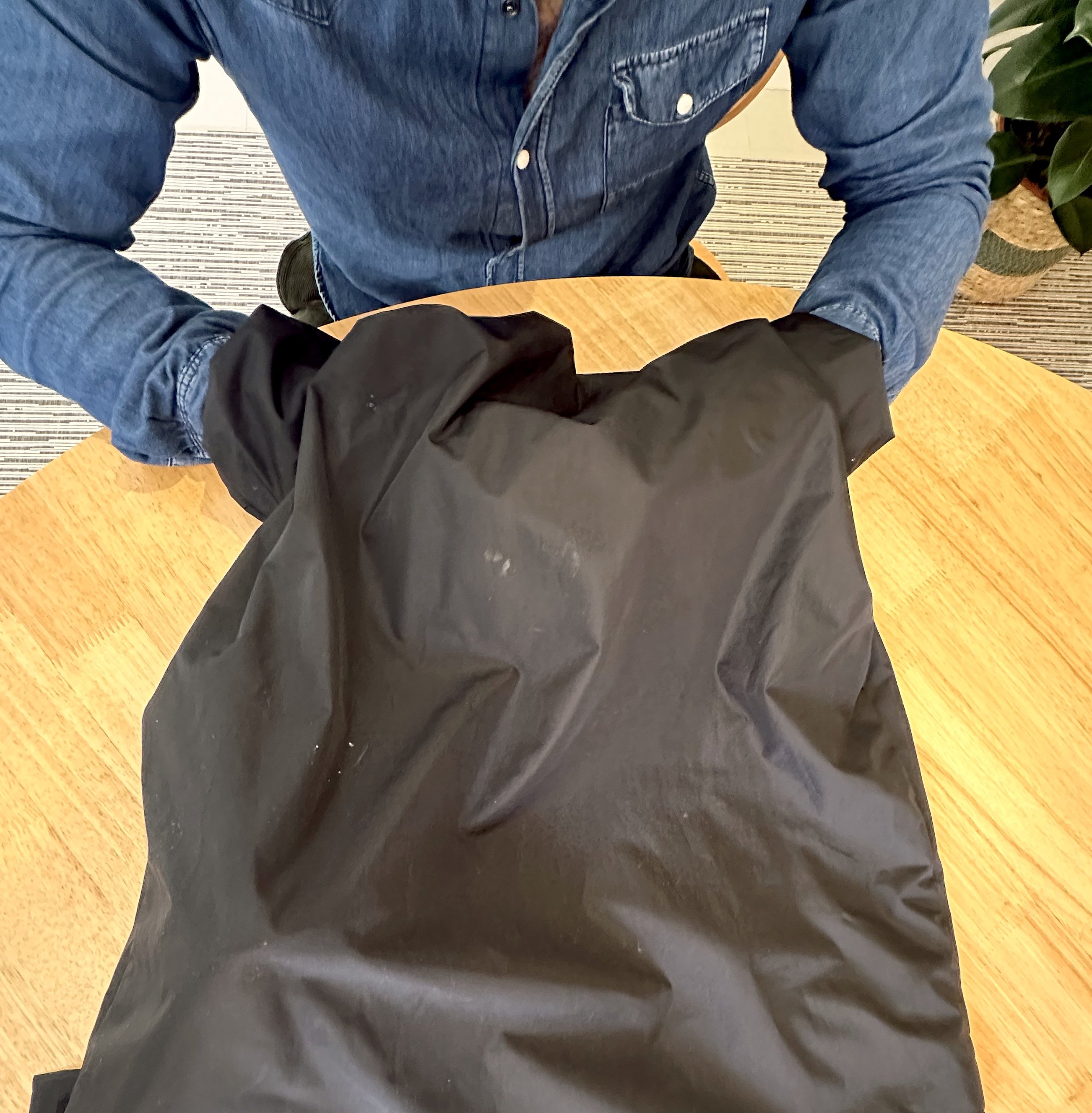 A person in a blue denim shirt sitting at a wooden table, handling a black plastic bag.