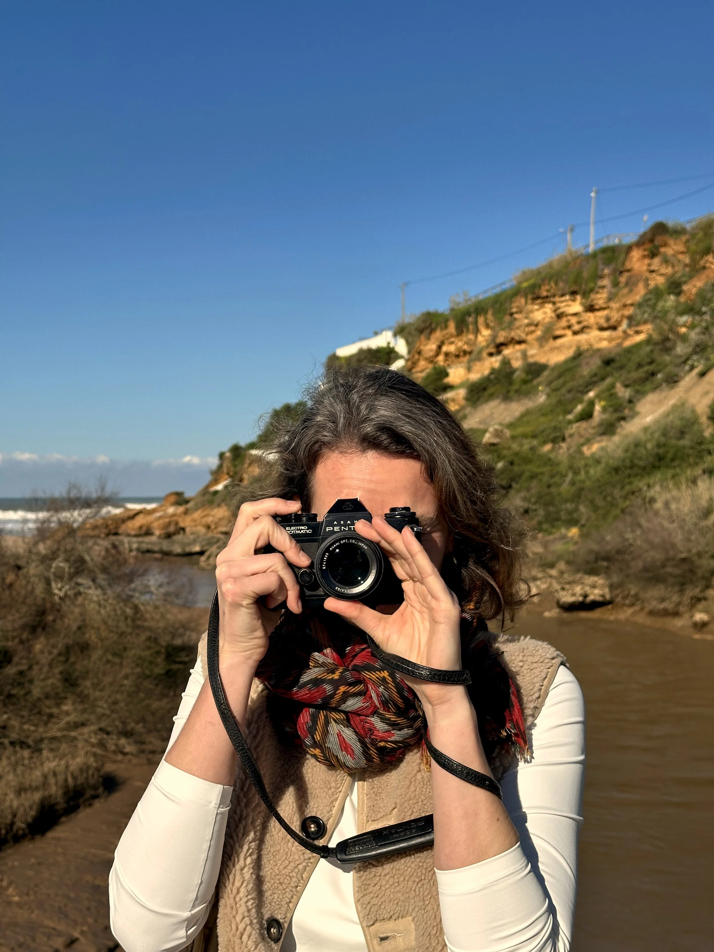 A woman with wavy hair taking a photograph with a vintage camera outdoors near a stream, with a hill and a house in the background under a clear blue sky.