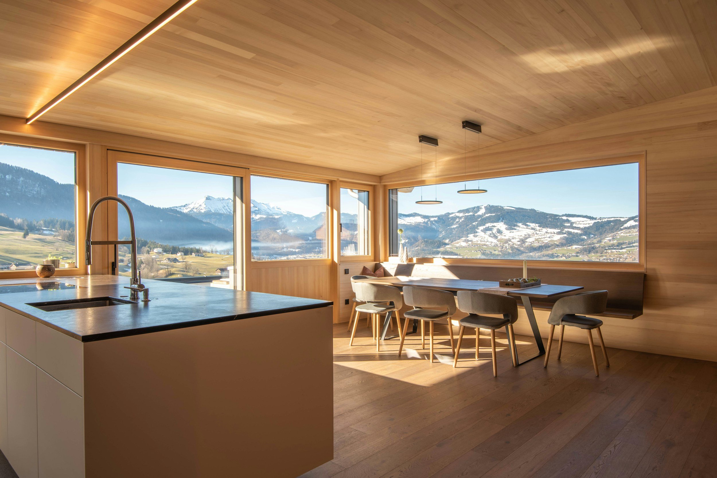 Modern kitchen with large windows showcasing mountain and valley view, wooden interior, kitchen island with sink, dining table with six chairs, minimalistic lighting fixtures