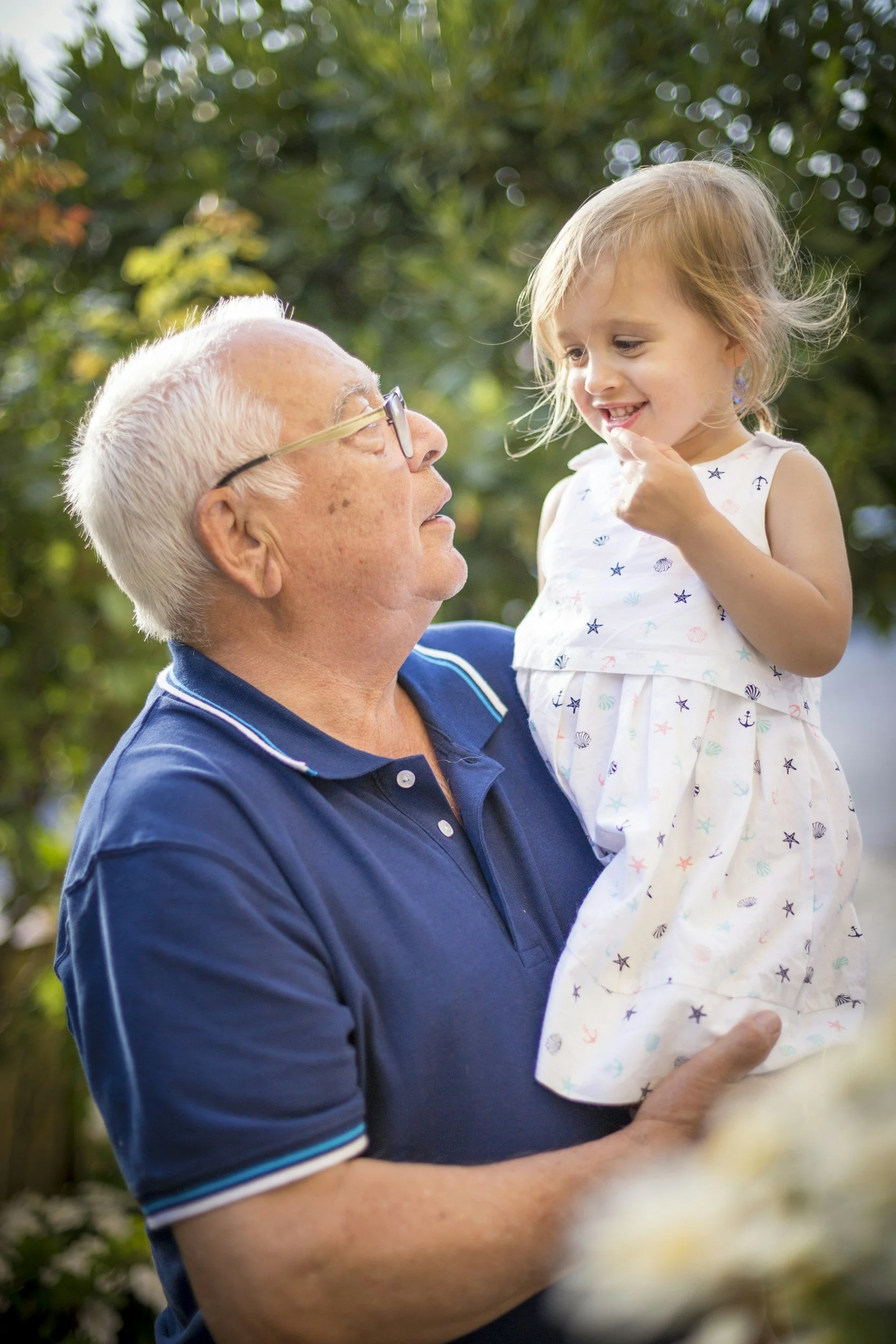 An older adult man holding a young girl in his arms outdoors, smiling and looking at her with greenery in the background.