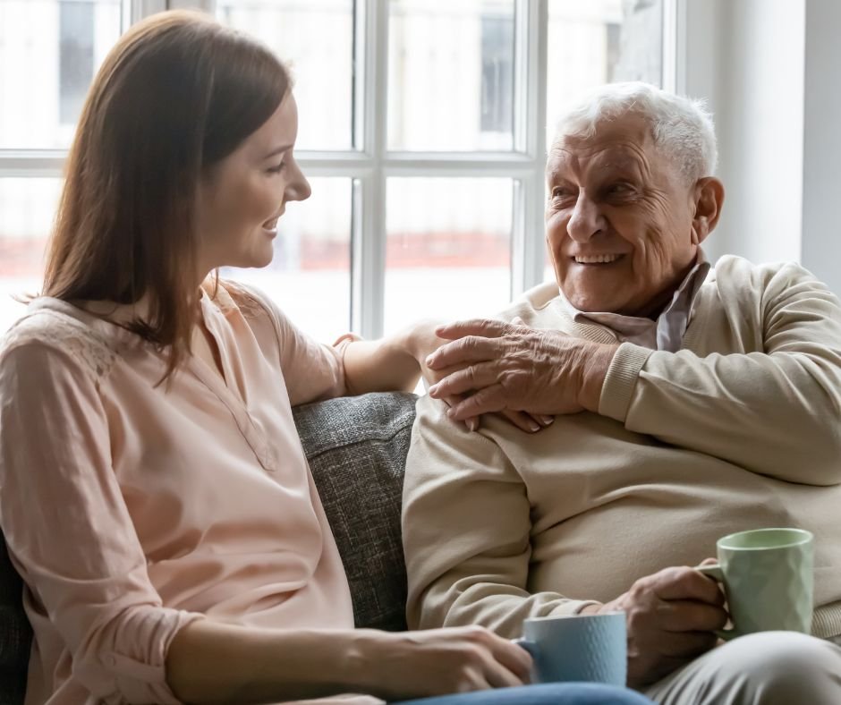 A young woman and an elderly man sitting together on a couch near a window, smiling and enjoying a conversation, each holding a mug.