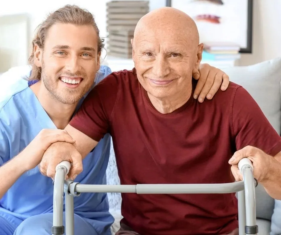 A young male nurse or caregiver with long hair and a beard smiling, assisting an elderly man with a walker, both smiling and sitting together in a bright room.