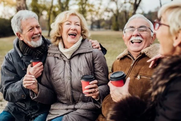 Four elderly people outdoors, smiling and laughing together while holding coffee cups.