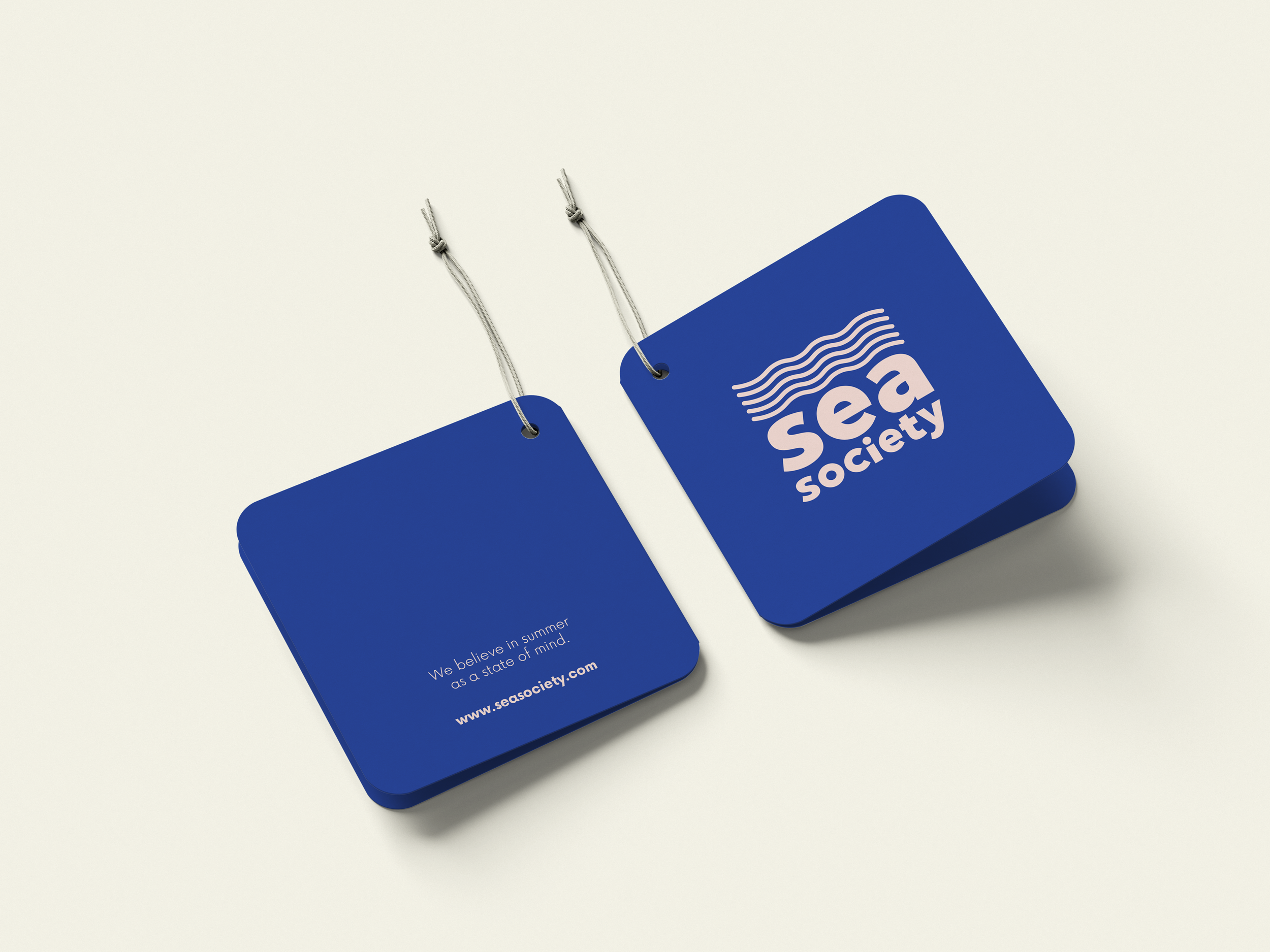 Dos etiquetas azules con cuerda, una con el logo de Sea Society y otra con la frase 'We believe in summer as a state of mind' y la página web www.seasociety.com.