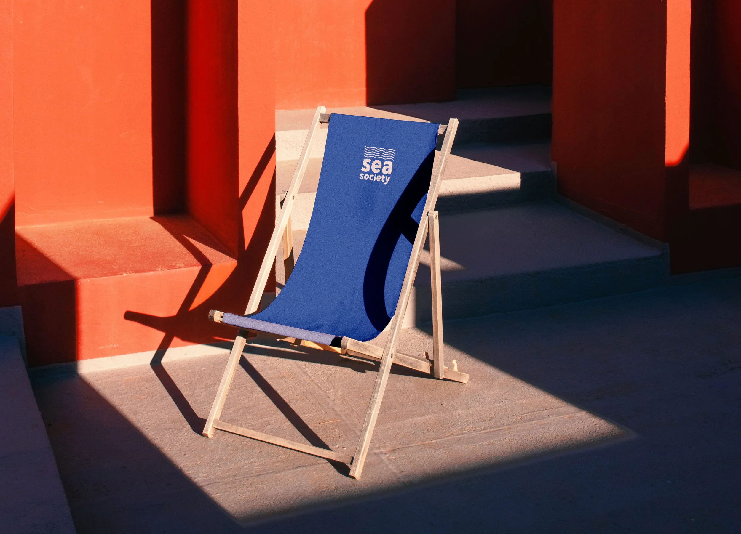 Silla de playa azul con logotipo de 'Sea Society' en un entorno con paredes rojas y sombras, cerca de unas escaleras de concreto.