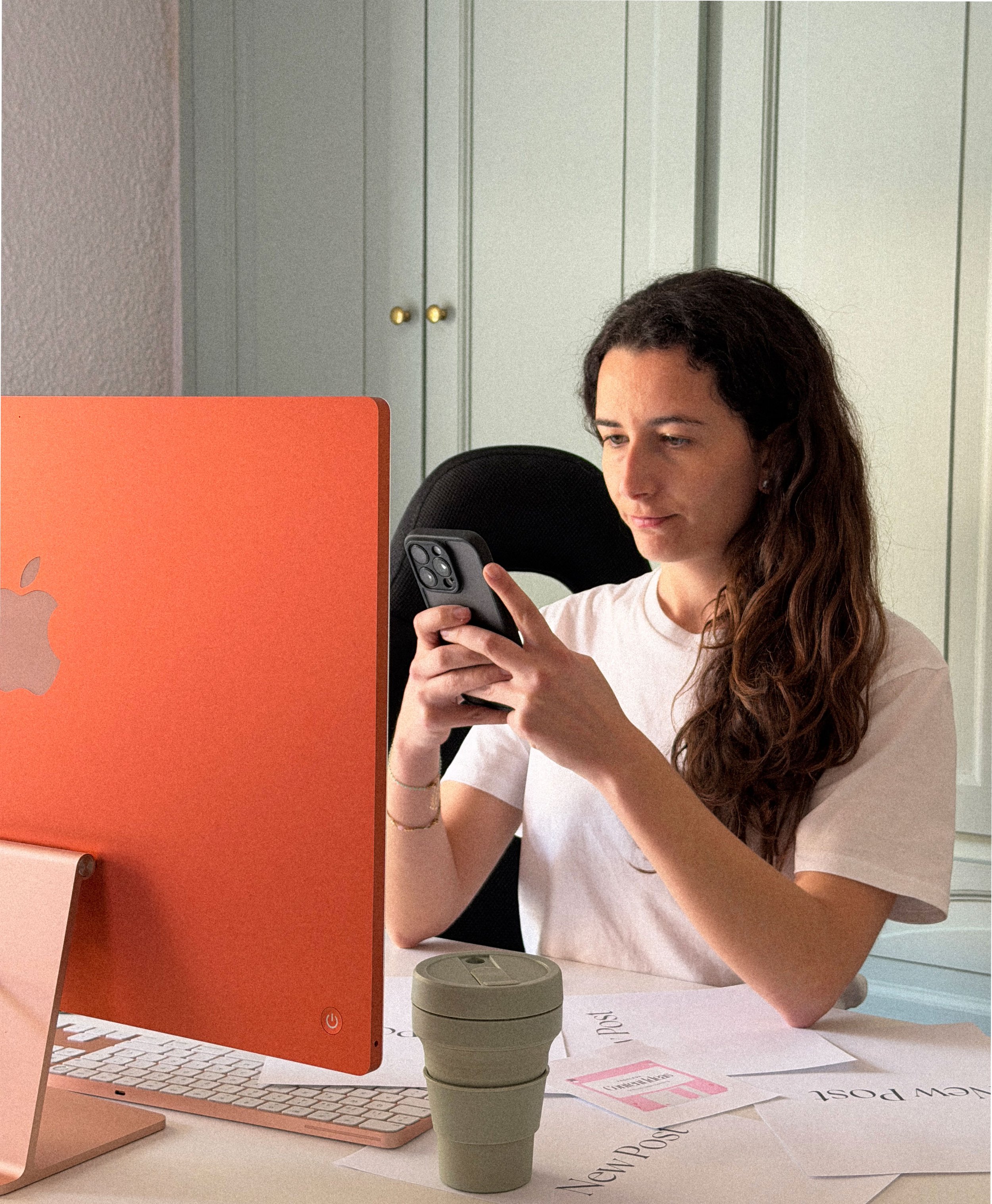 Mujer sentada en un escritorio, usando un teléfono móvil, con una computadora y documentos sobre la mesa.