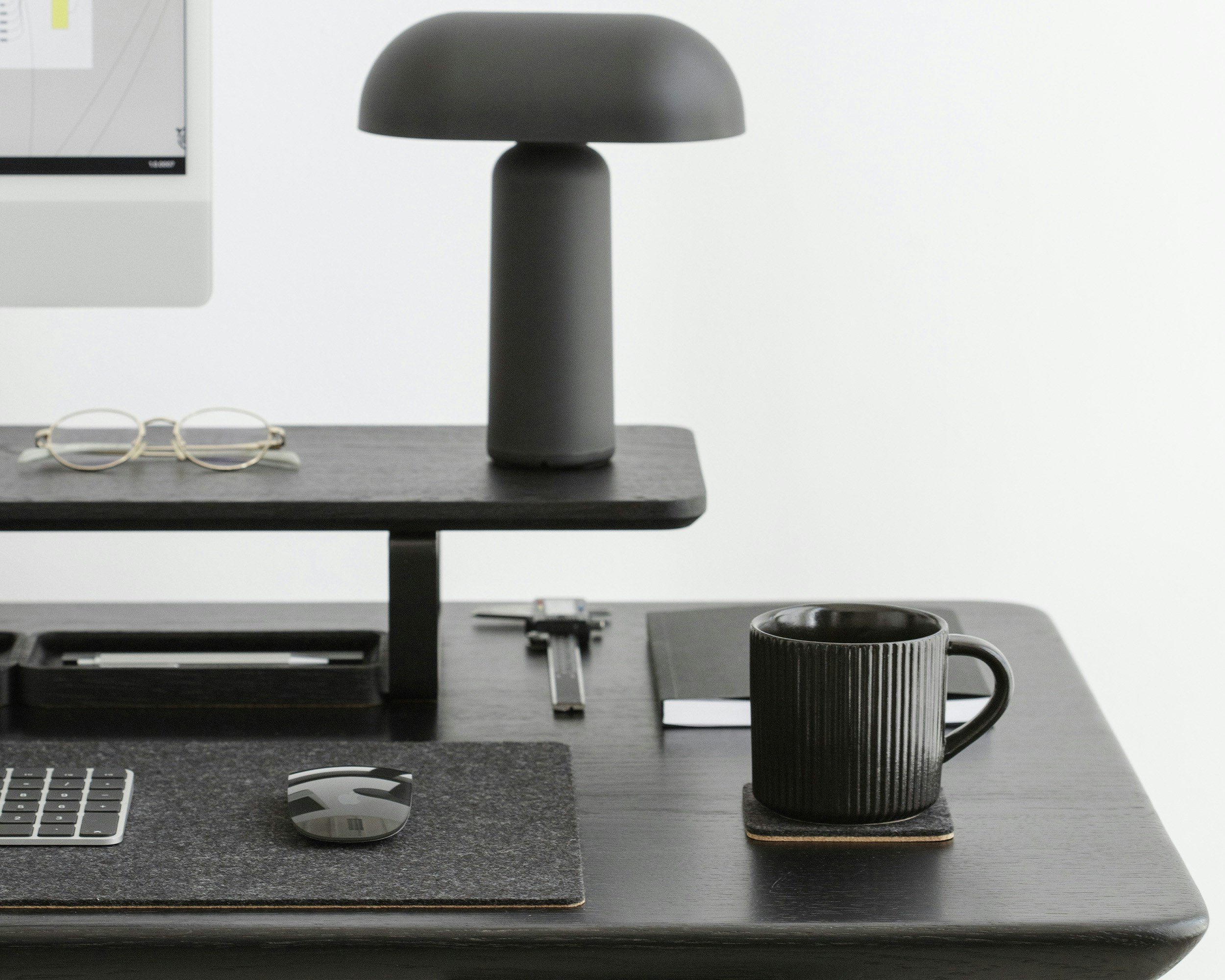 A modern black office desk setup with a black coffee mug on a coaster, a computer mouse and keyboard, glasses, a stapler, a small ruler, a notebook, a table lamp, and a computer monitor in a minimalist workspace.