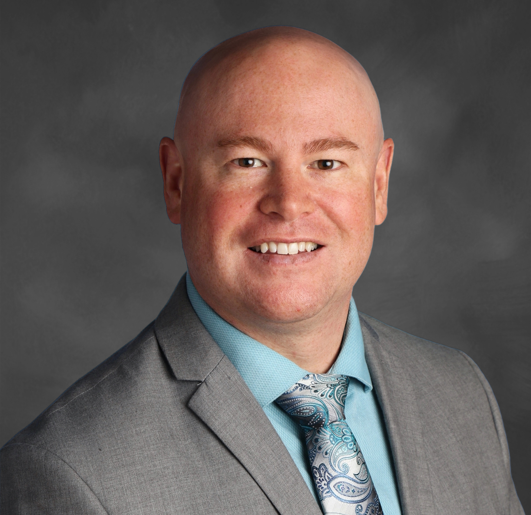 A professional headshot of a man with a bald head, no facial hair, wearing a gray suit, light blue shirt, and a patterned blue and gray tie, smiling against a dark gray background.