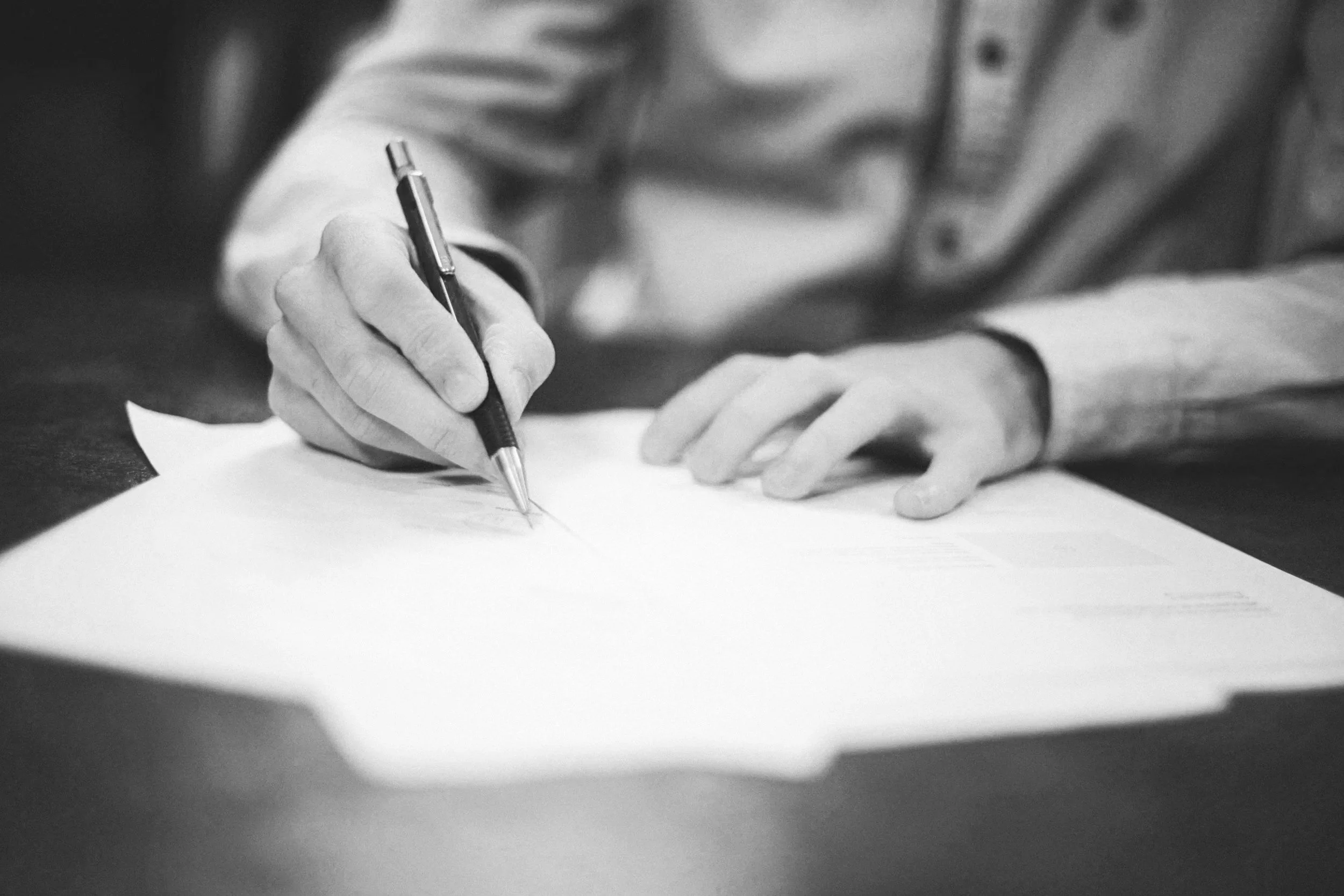 Black and white image of a person in a long-sleeve shirt writing on a piece of paper with a pen, resting their other hand on the paper.